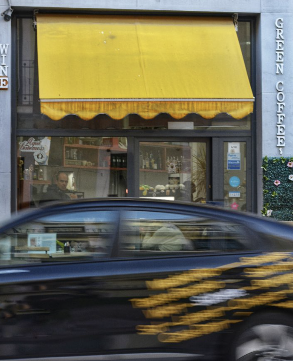Blurred black car passing in front of a coffee shop with a yellow awning outside. Inside the shop, a person is visible through the window, sitting at a counter with shelves of items behind them.