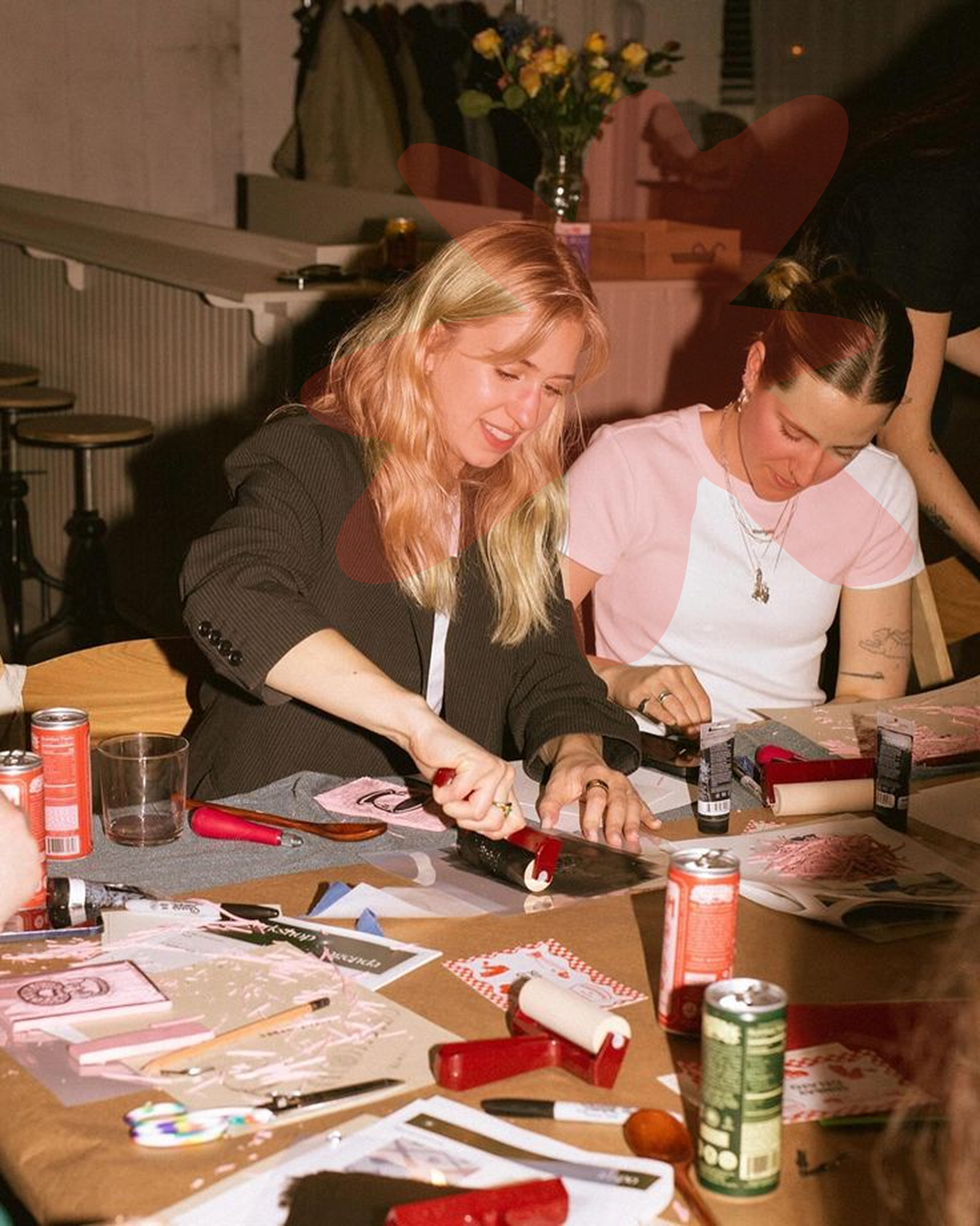 Two women sitting at a table opening a gift, surrounded by cans, paper, scissors, and crafting supplies, with a woman in black serving as a photographer or assistant, in a cozy indoor space with a vase of yellow flowers in the background.