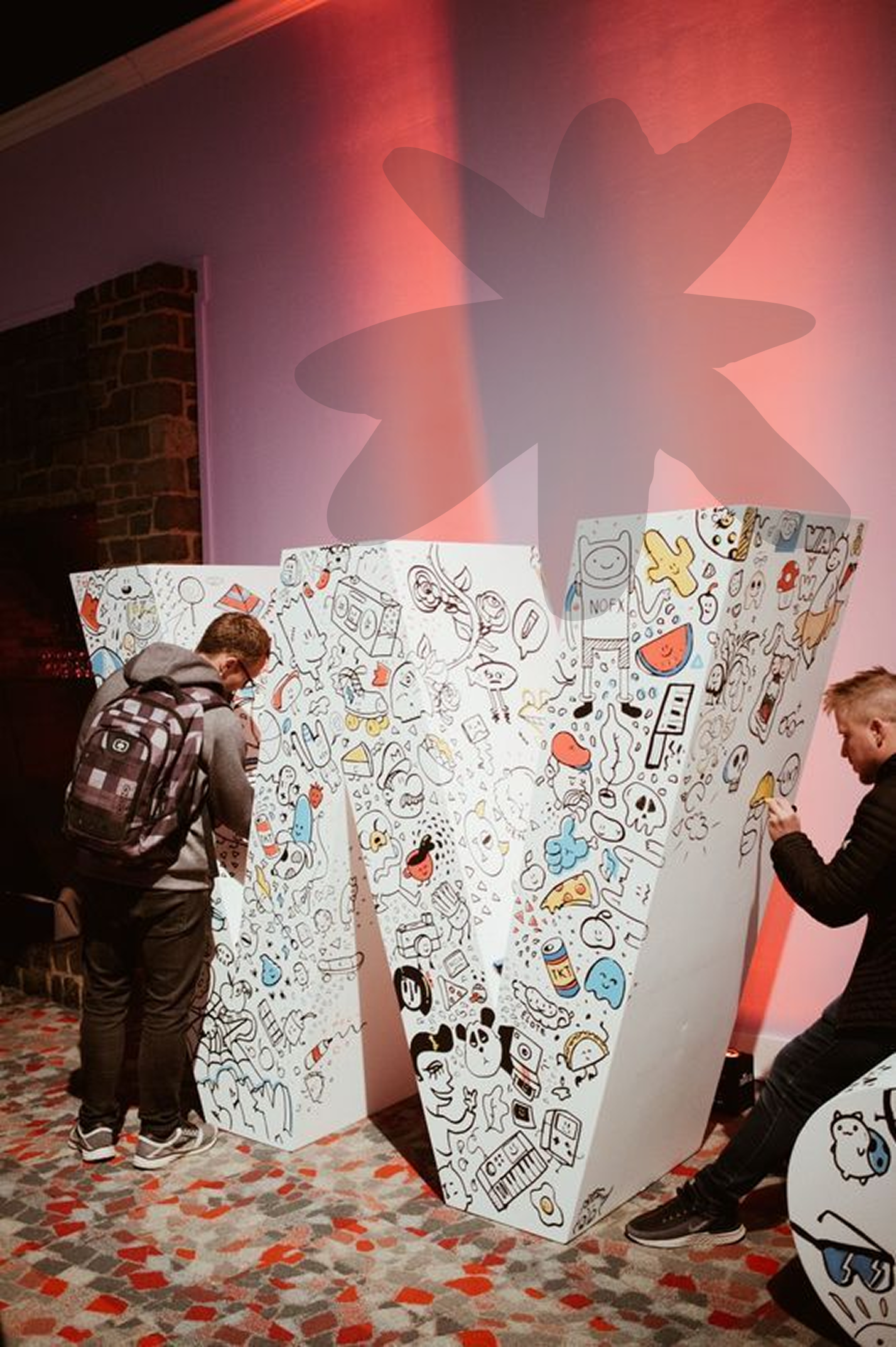 Two people drawing with markers on large white panels decorated with doodles of various cartoon characters and objects, in a room with a patterned carpet and a brick wall.