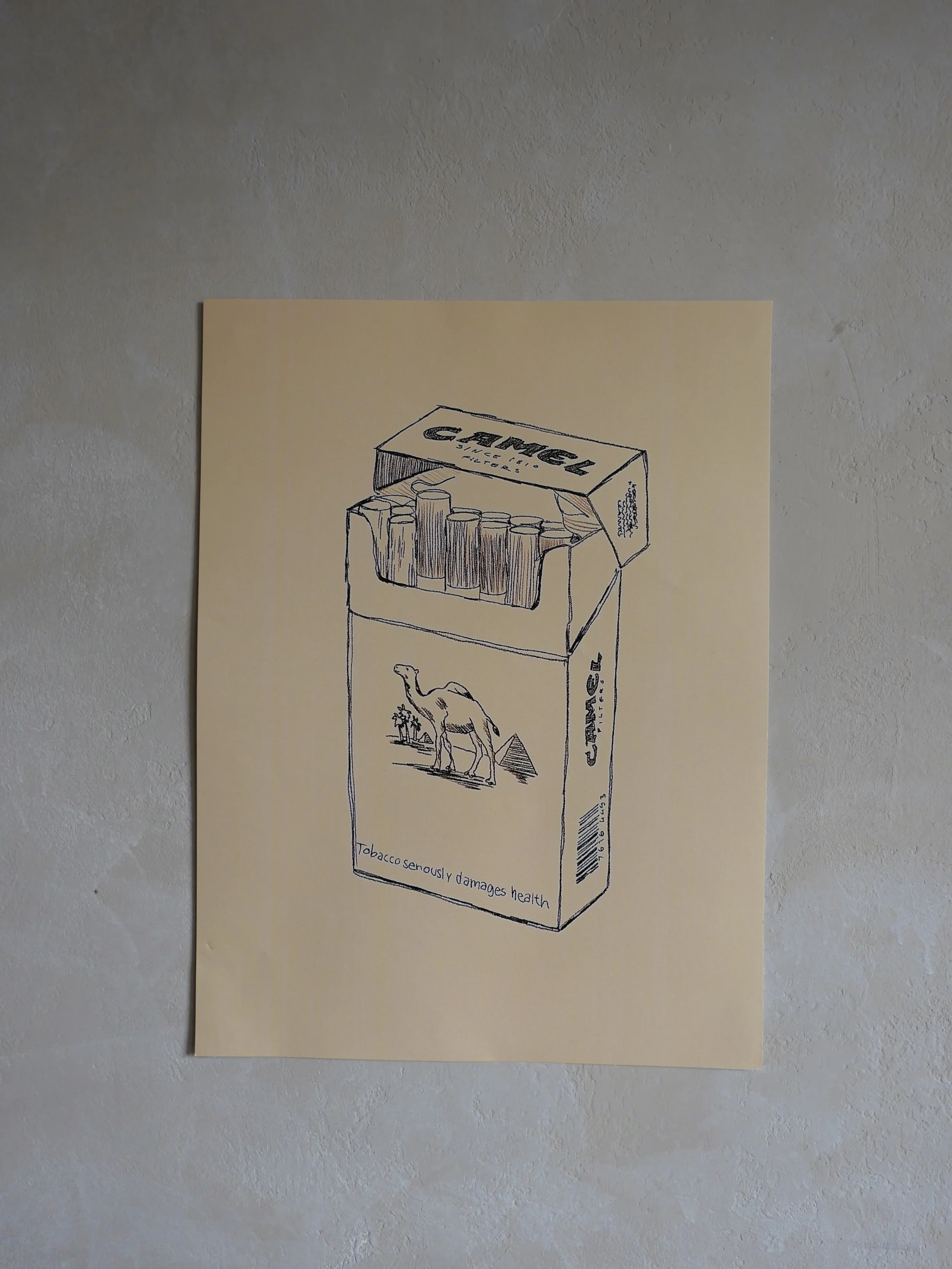 Hand-drawn sketch of a cigarette pack labeled "CAMLCE" with a camel illustration and a warning that says "Tobacco seriously damages health" on a beige paper taped to a light-colored wall.