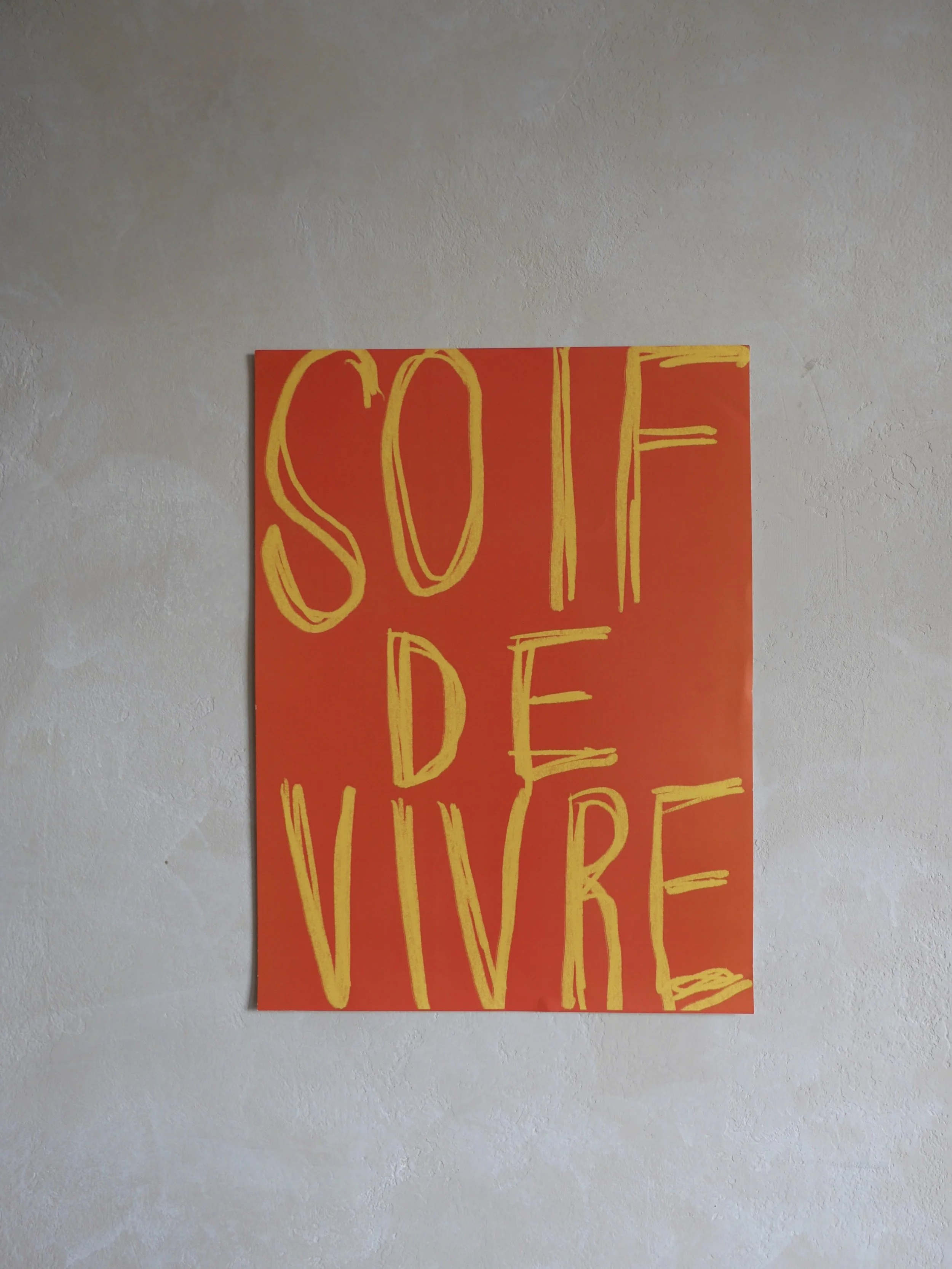 Red poster with yellow hand-drawn French text 'SOUFFRE DE VIVRE' on a textured white wall.