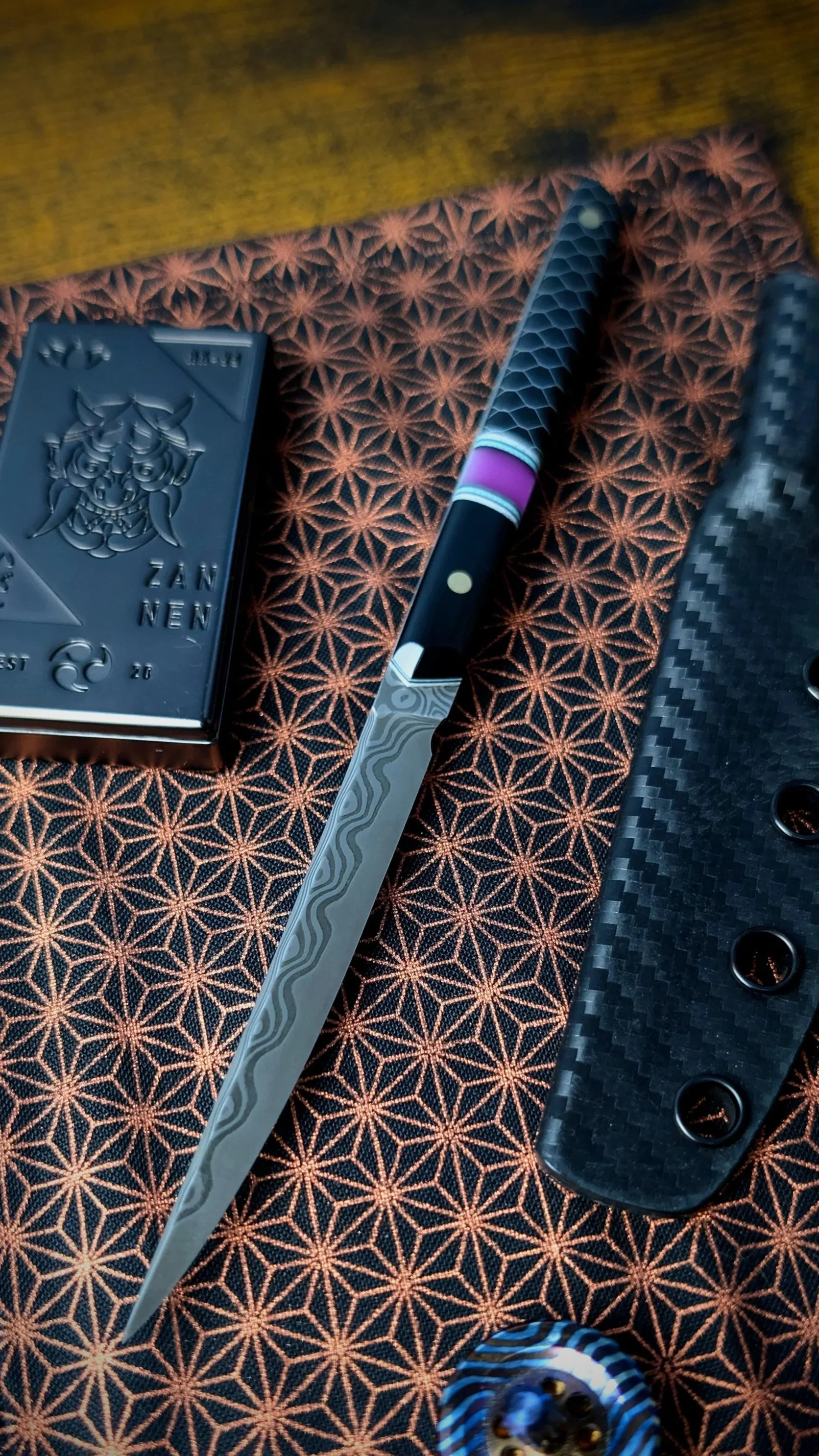 A close-up of a Japanese-style folding knife with a patterned blade, resting on a fabric surface with a star-like pattern. A black knife handle with circular cutouts made of carbon fiber is partly visible on the right, alongside a small blue and white striped bowl with golden decorative balls.