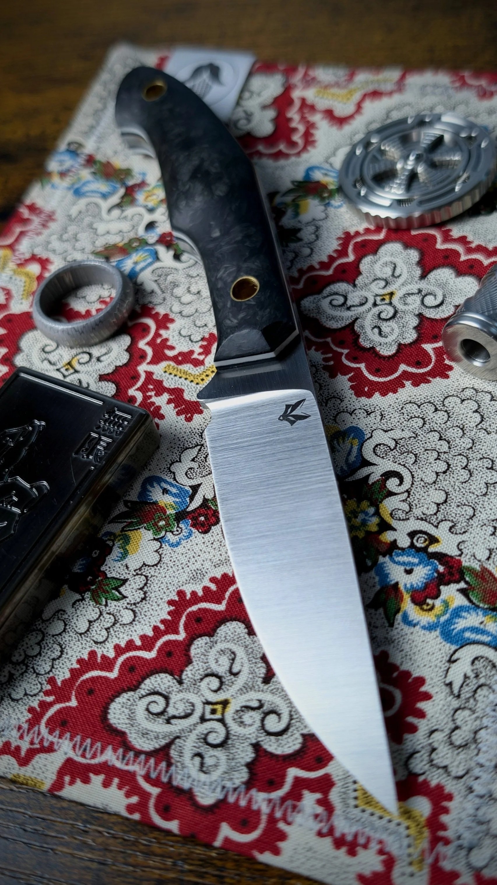 Close-up of a knife with a black handle and gray blade placed on a decorative red, white, and blue embroidered cloth with floral patterns, along with other metallic objects.