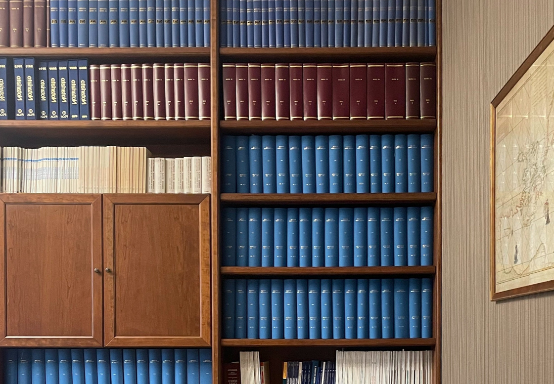 Scaffale con libri rilegati in blu, rosso e bianco, parte di una libreria in legno con mappa appesa a destra.