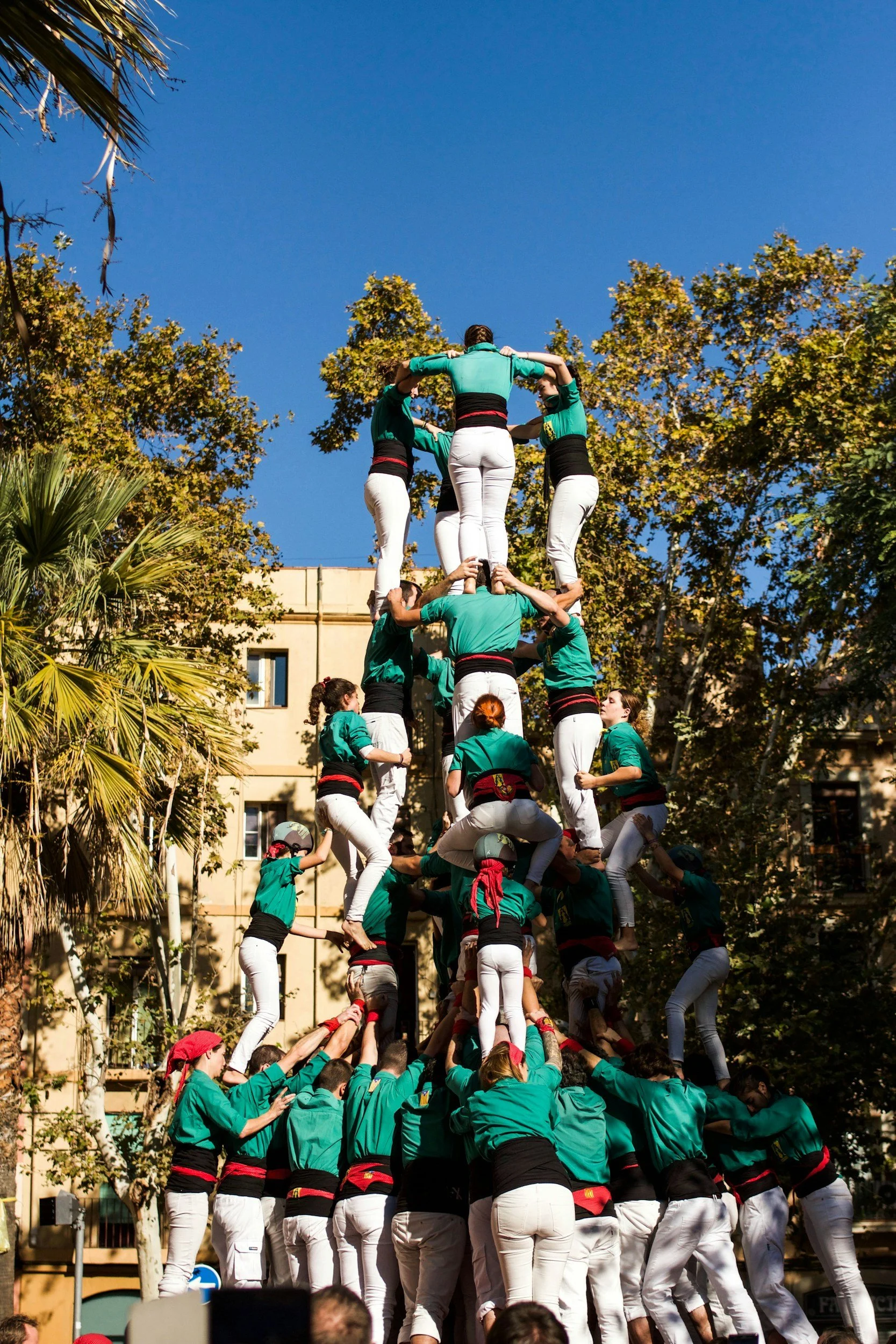 Human tower or "castell" being formed, with people in green shirts and white pants, outdoors against a backdrop of trees and buildings.