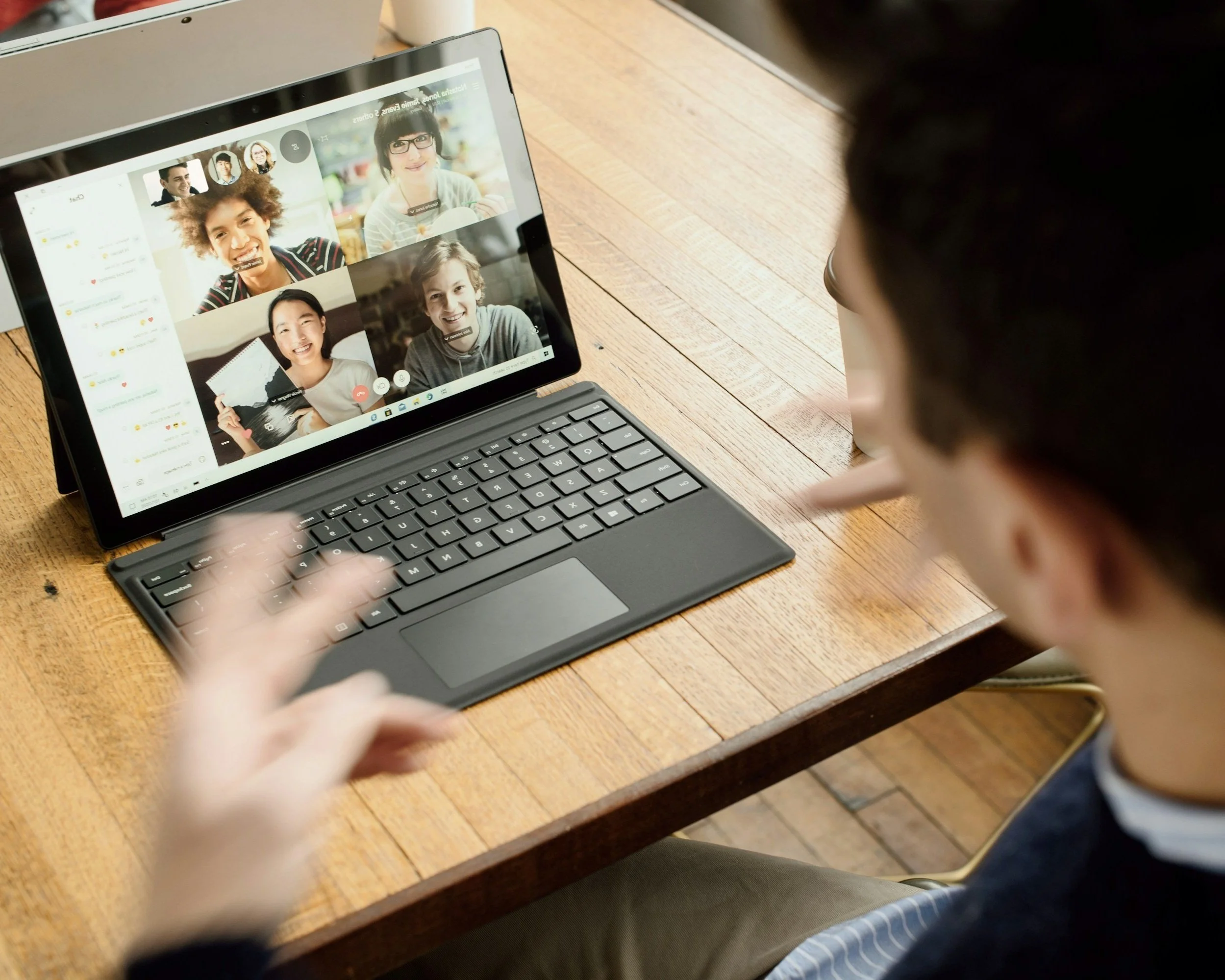 Person participating in a video conference on a tablet, showing multiple people on the screen, seated at a wooden table.