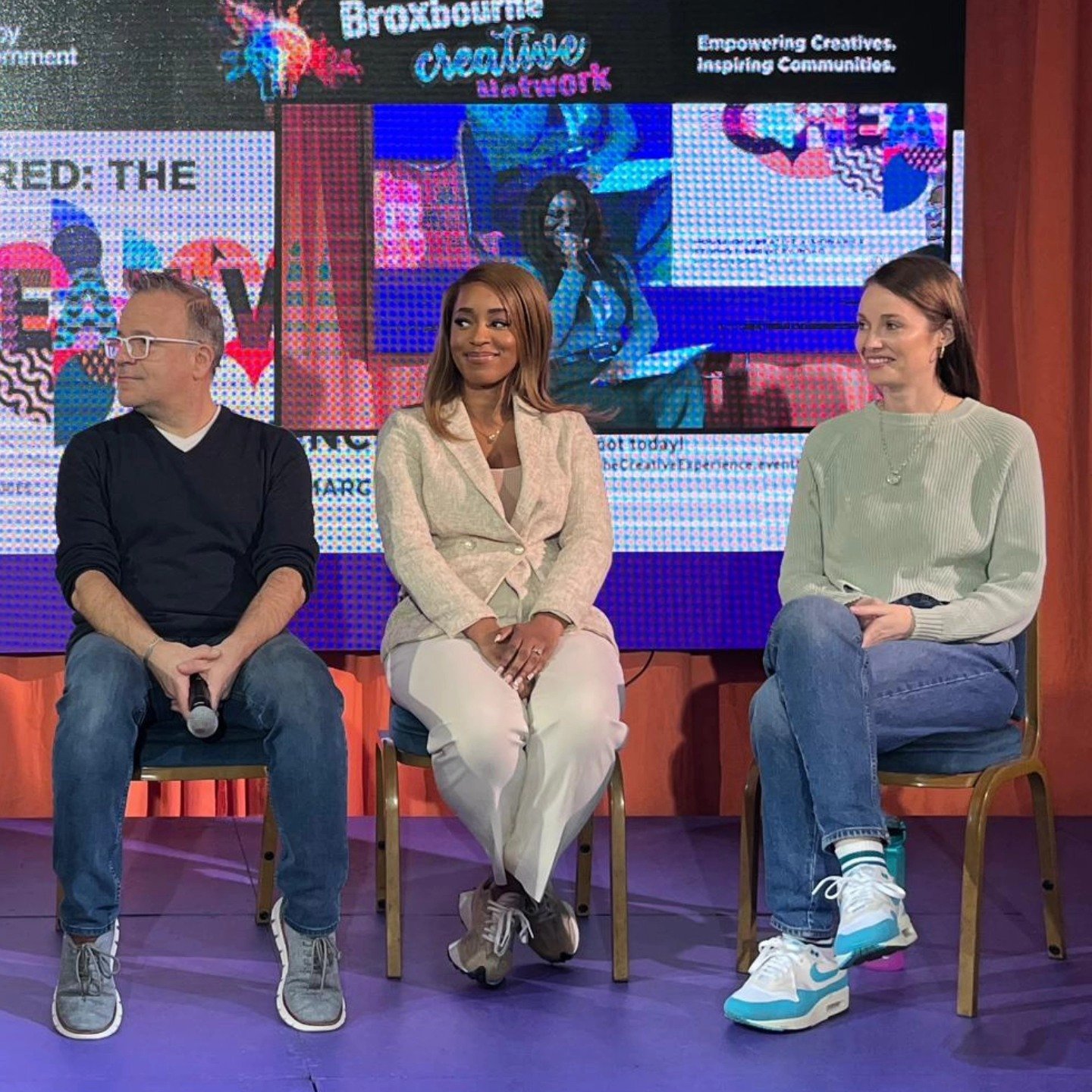 Three people seated on stage at the Broxbourne Creative Network event, with a background displaying digital screens.