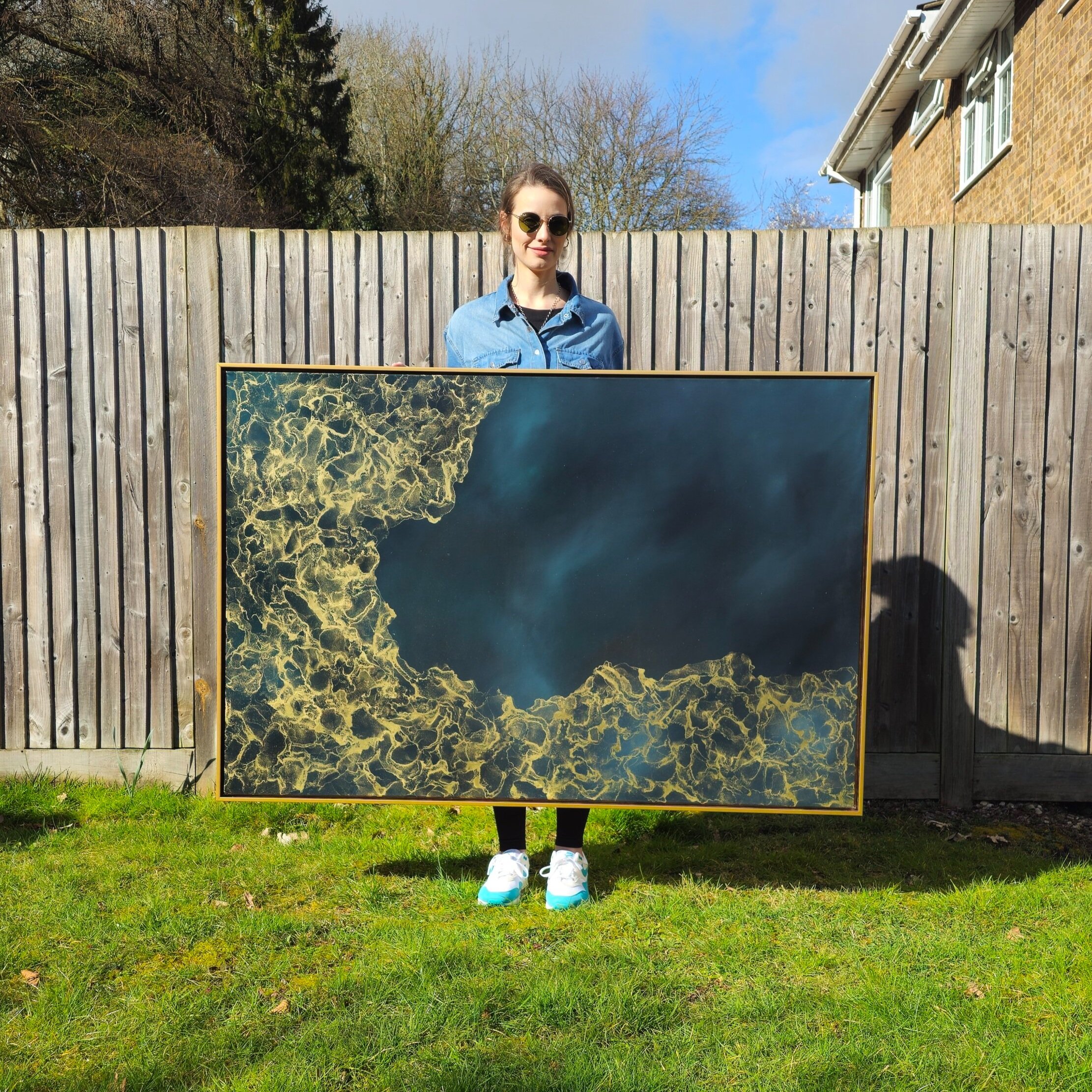 Person holding a large abstract painting with a golden pattern on a blue background standing outside against a wooden fence.