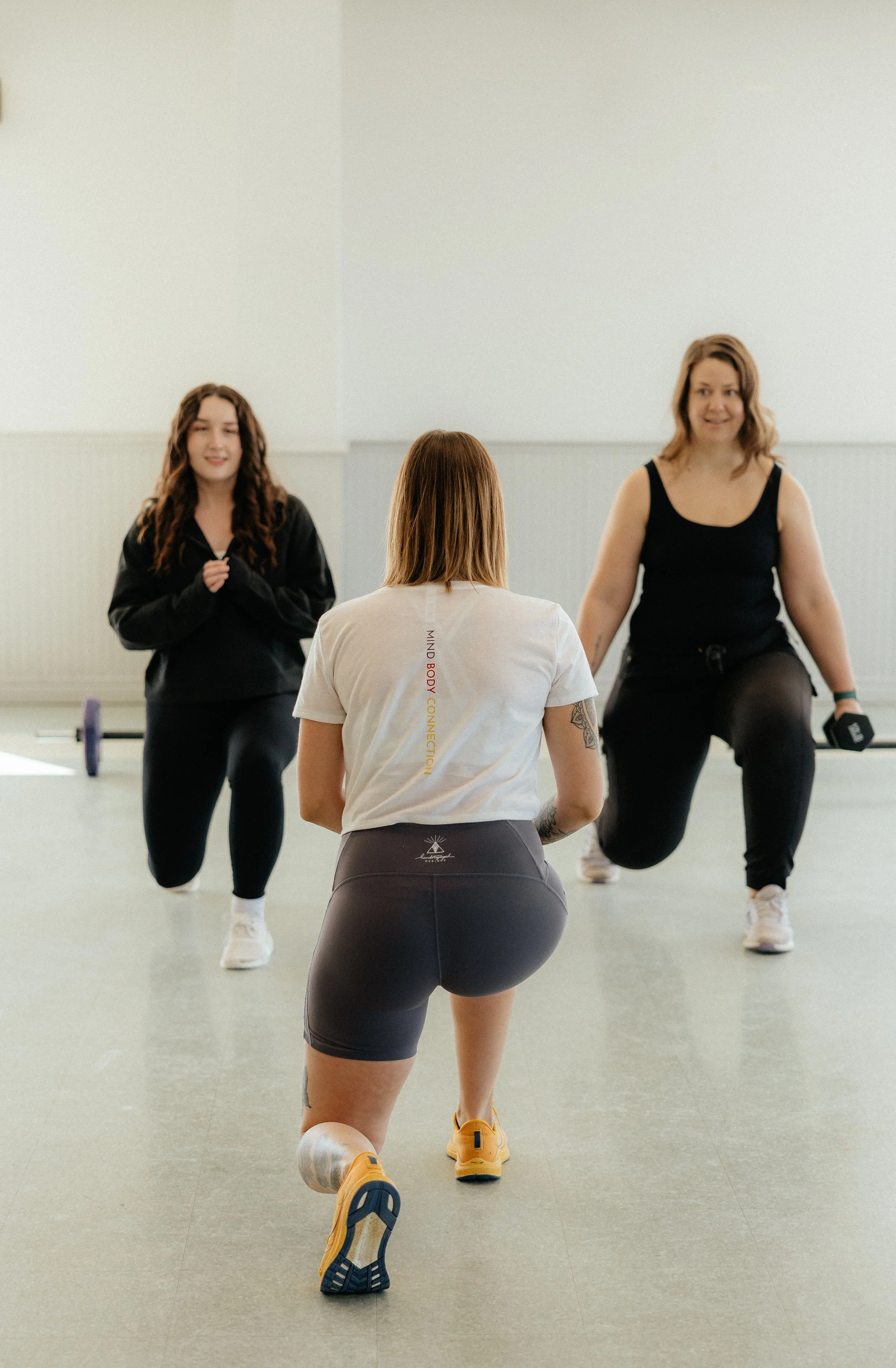 Three women are in a fitness class. One woman, seen from the back, is kneeling on one knee and facing the other two women. The other two women are standing and holding weights.