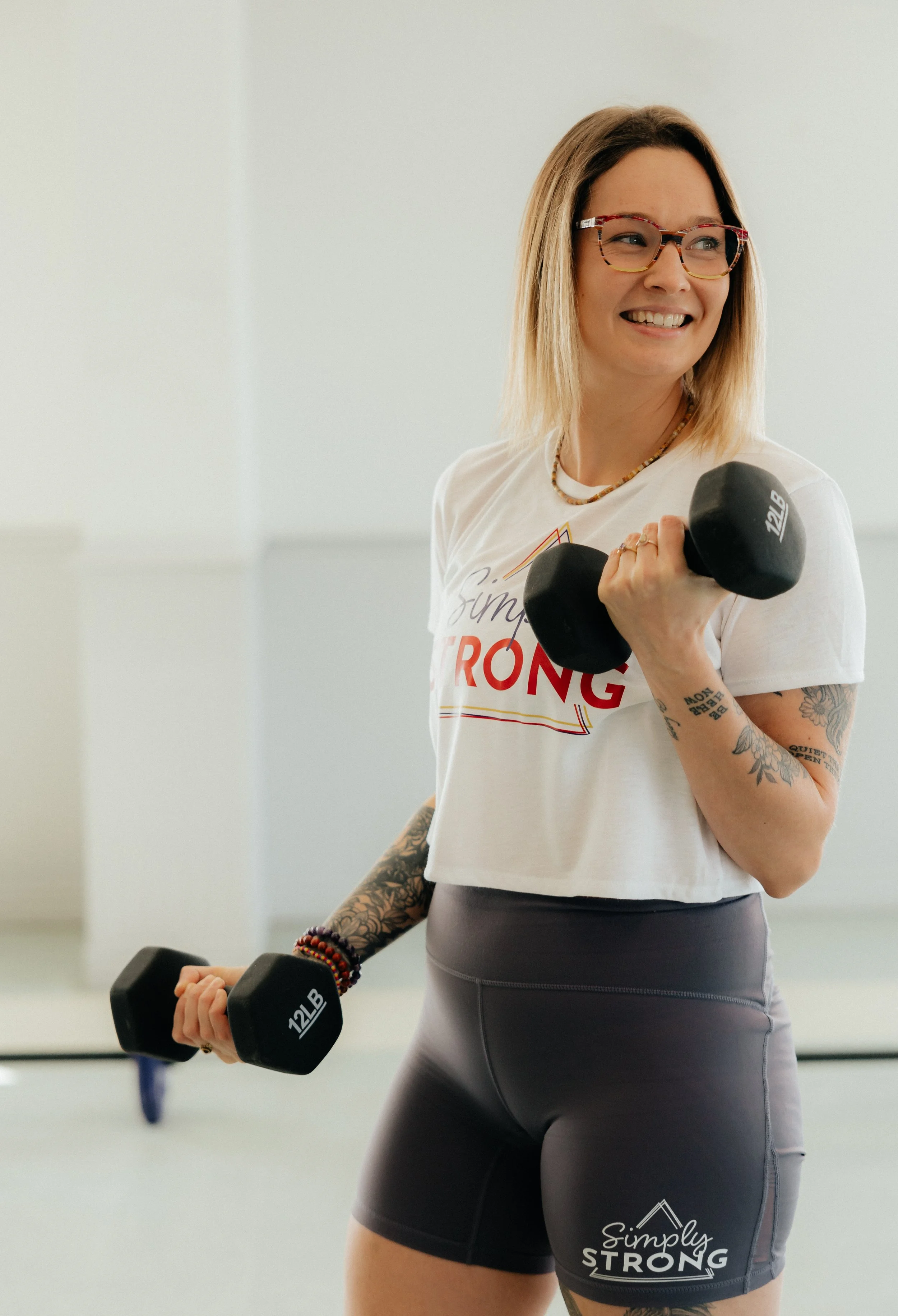 A smiling woman with glasses and tattoos exercising with dumbbells in a gym, wearing a white t-shirt and black shorts.