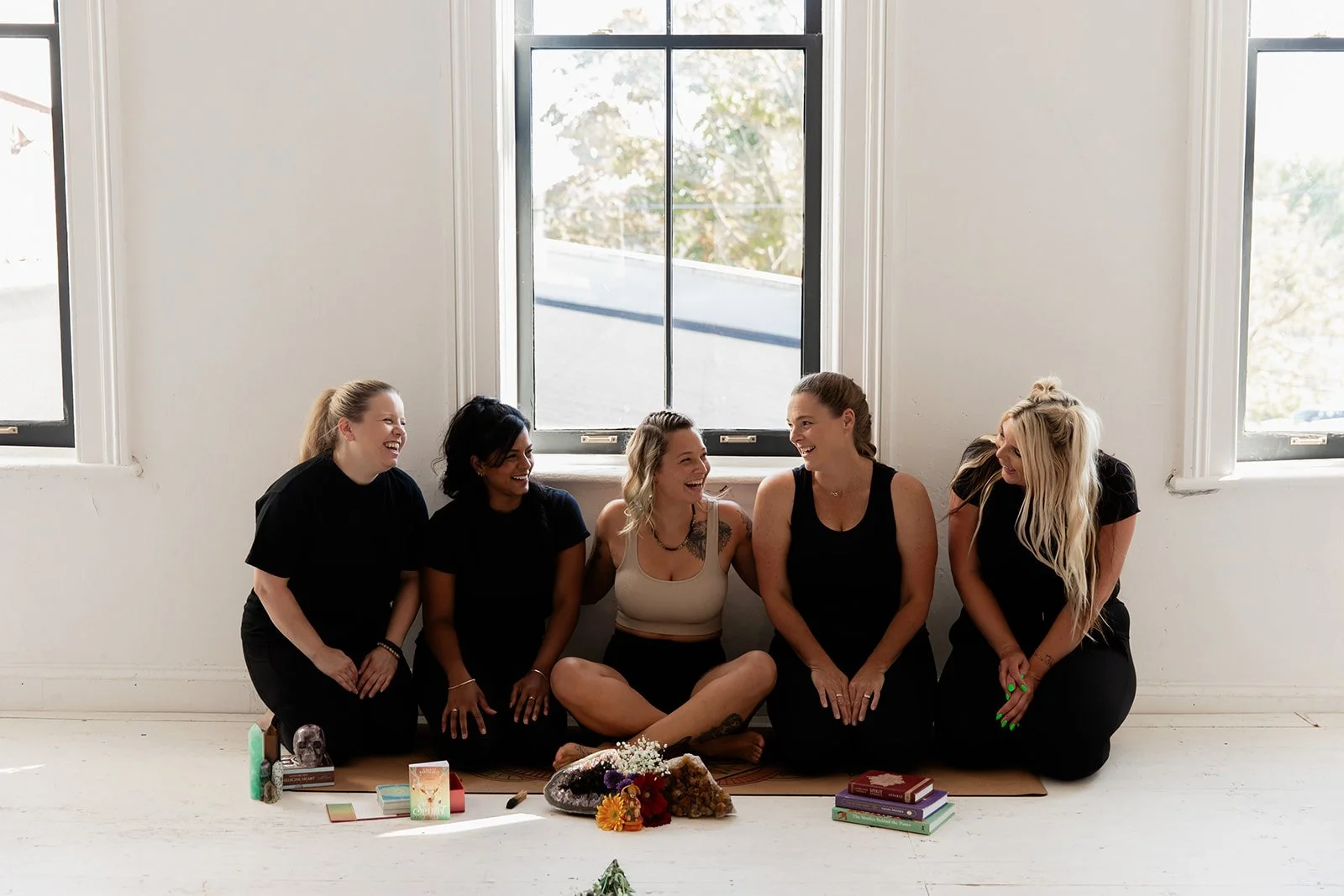 Five women sitting on the floor in a bright room, smiling and laughing together with windows behind them, surrounded by books and crystals.