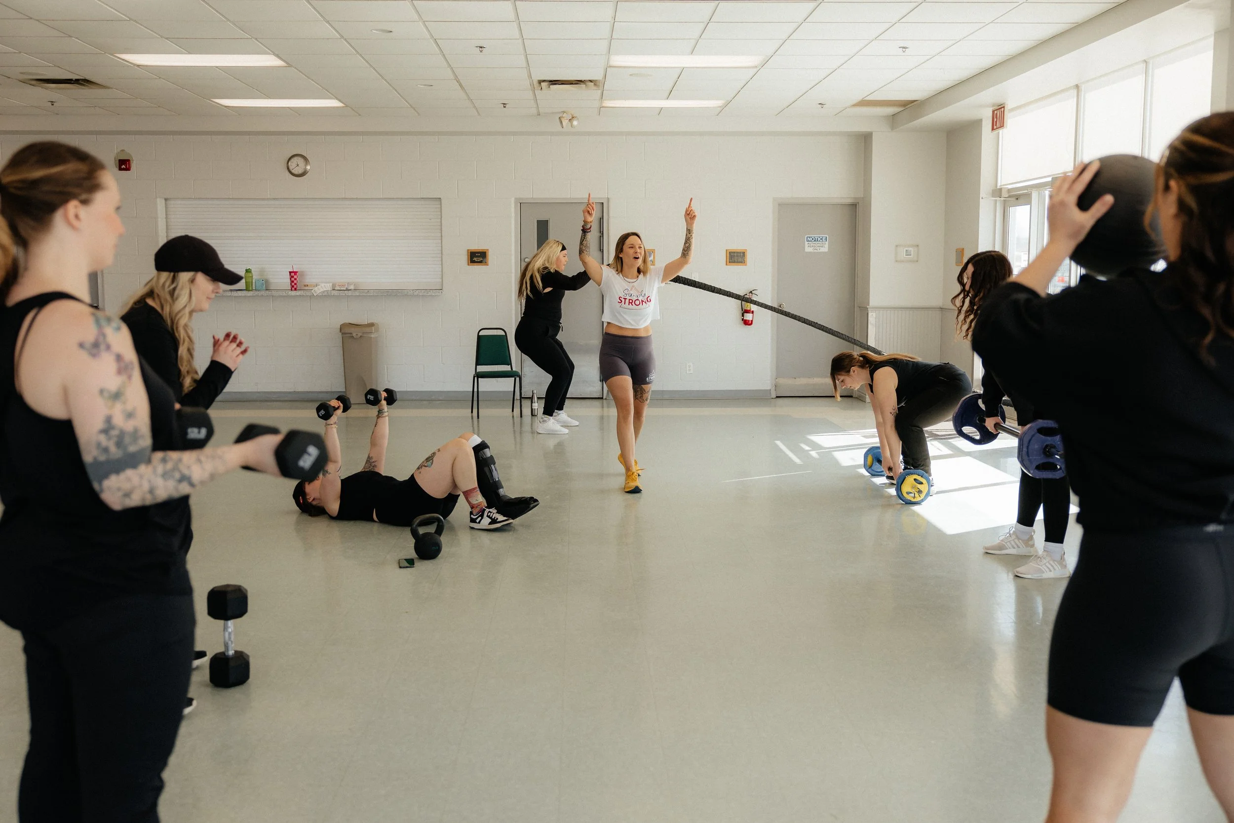 Group of women working out in a gym, some lifting weights and others doing exercises, with one woman instructing.