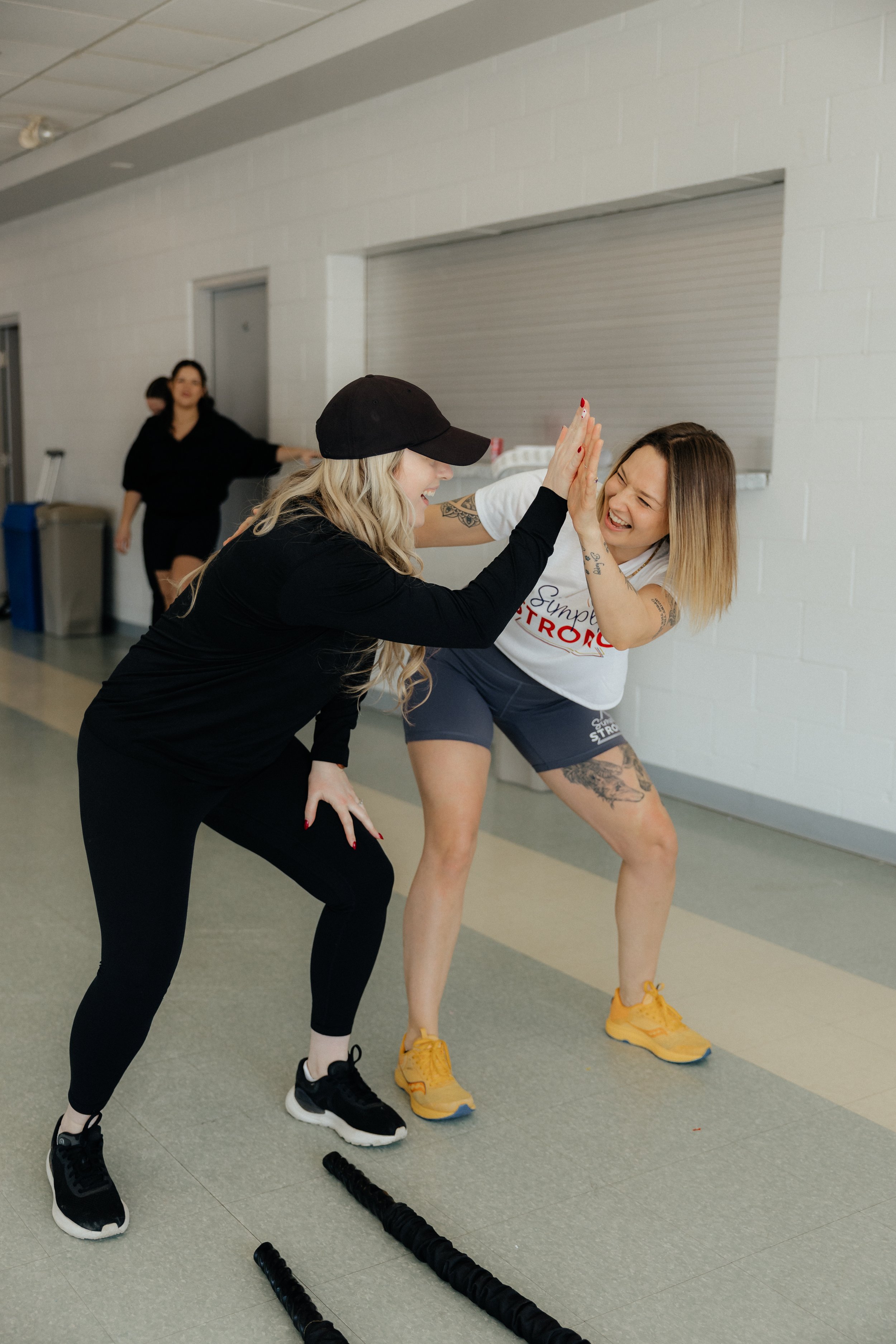 Two women in athletic clothing giving each other a high five inside a gym, with two people in the background and black agility sticks on the floor.