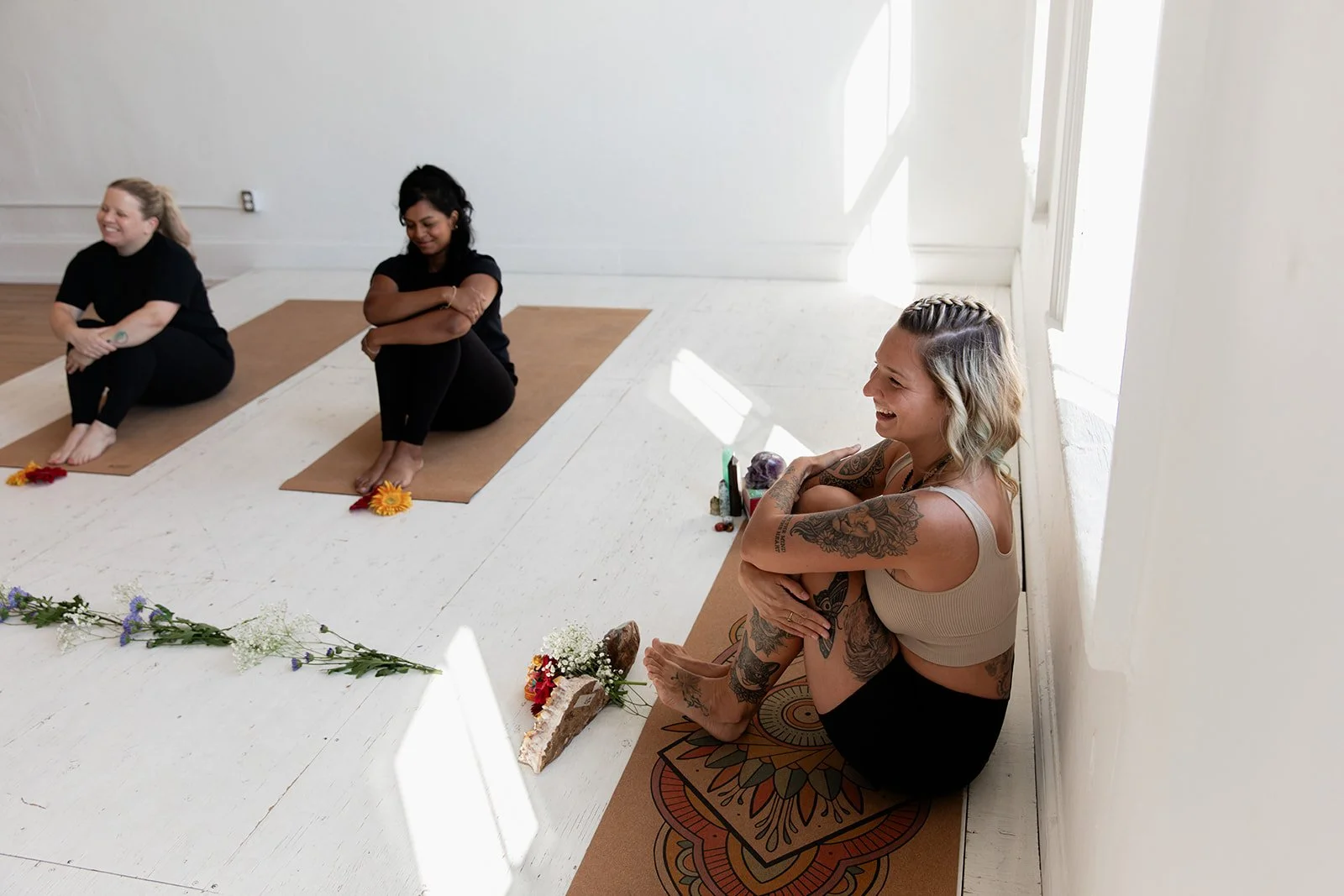 Three women sitting on yoga mats in a bright room, surrounded by flowers, smiling and relaxed.