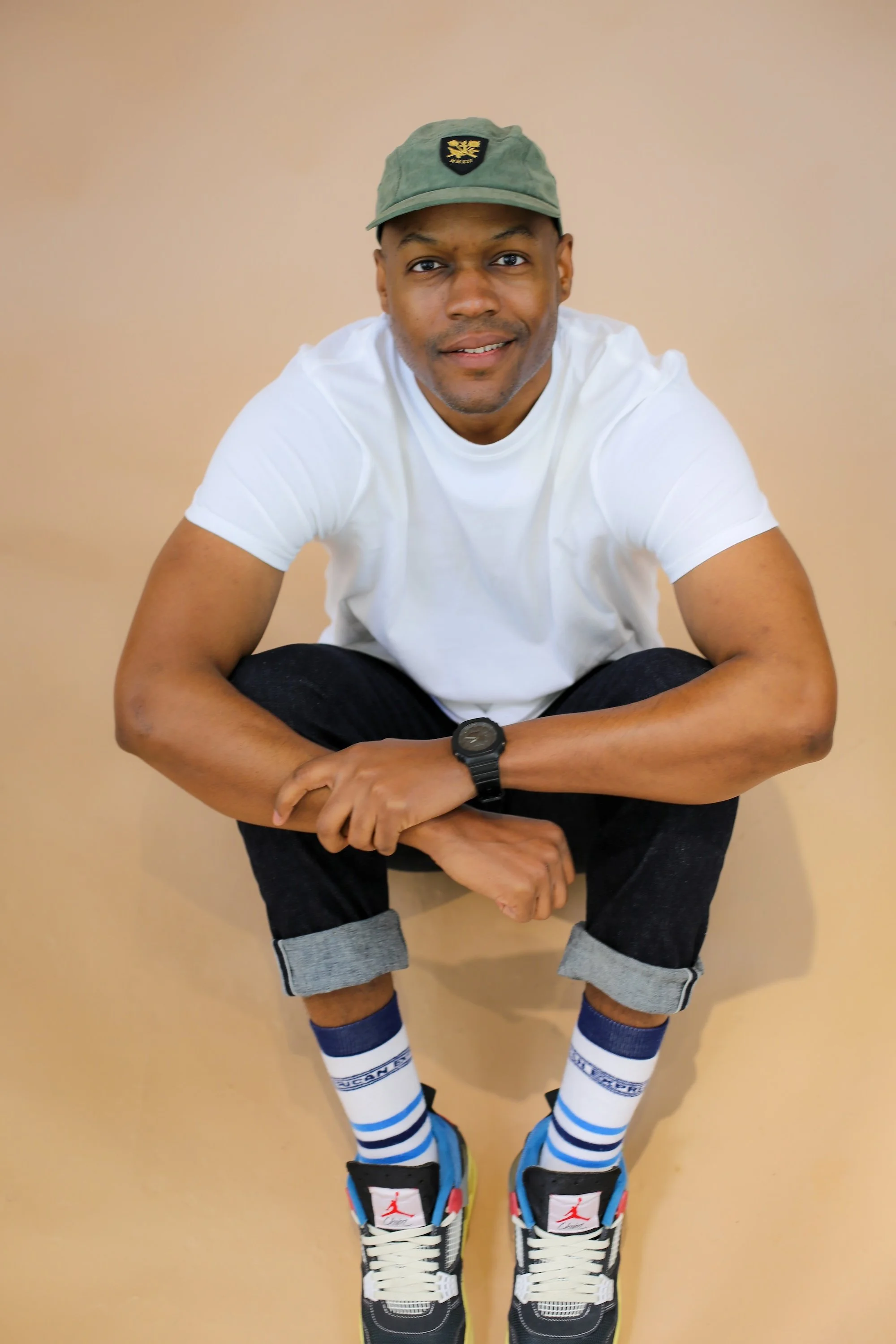 A young man in a white t-shirt and rolled-up black jeans crouching against a beige background, wearing a green cap, blue and white striped socks, and black and red sneakers.