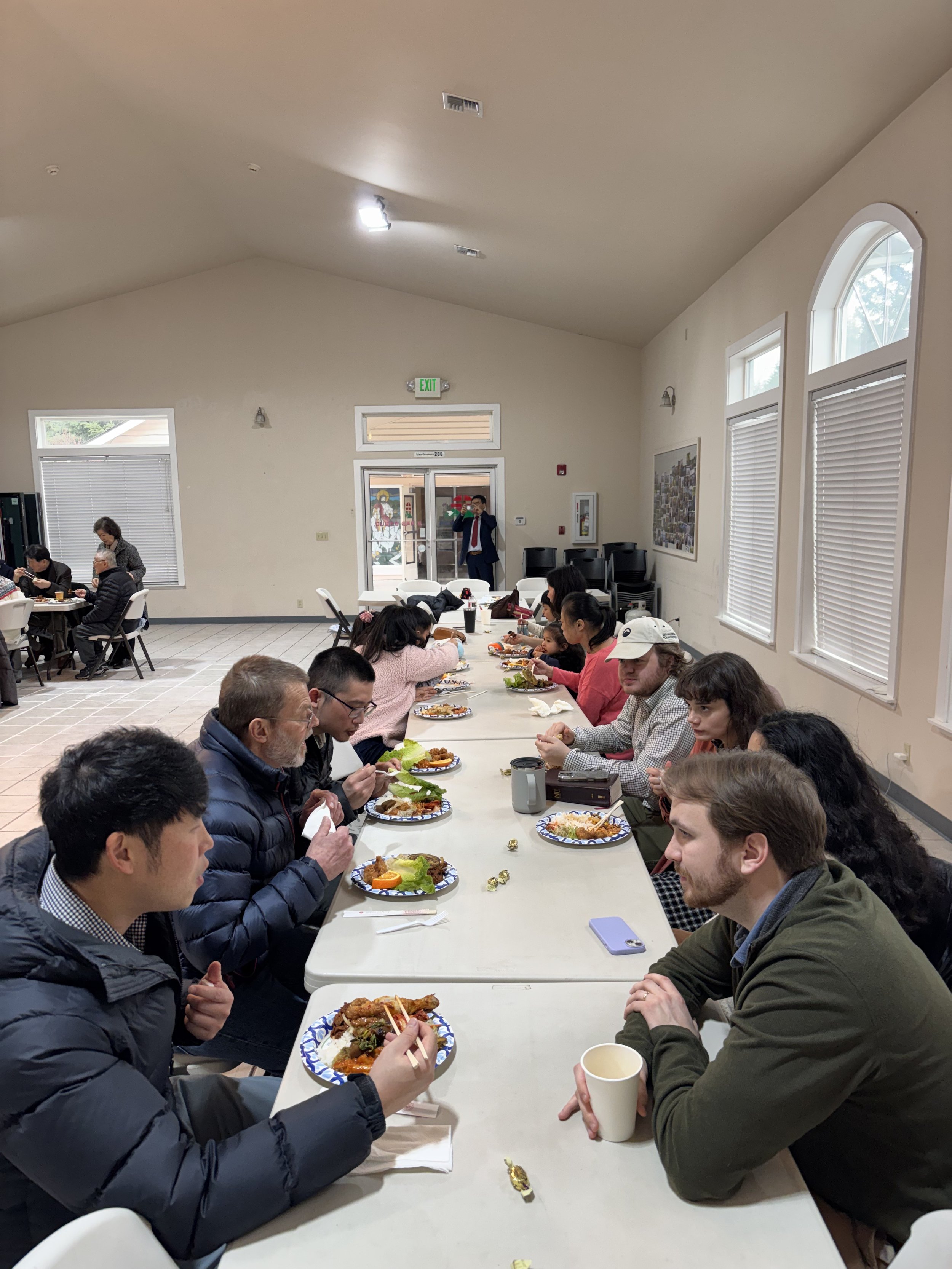 People sitting at a long table in a large room, eating a meal with plates of food in front of them.