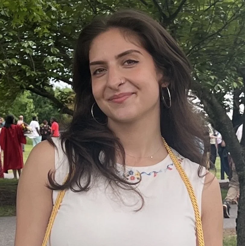 A young woman with dark hair, hoop earrings, and a nose ring smiling outdoors near a tree, with people in the background at a park or outdoor gathering.