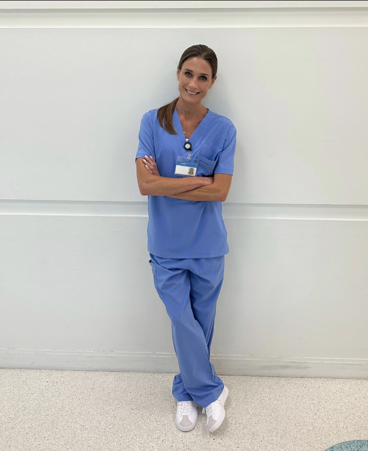 Person in blue scrubs standing and smiling against a white wall.