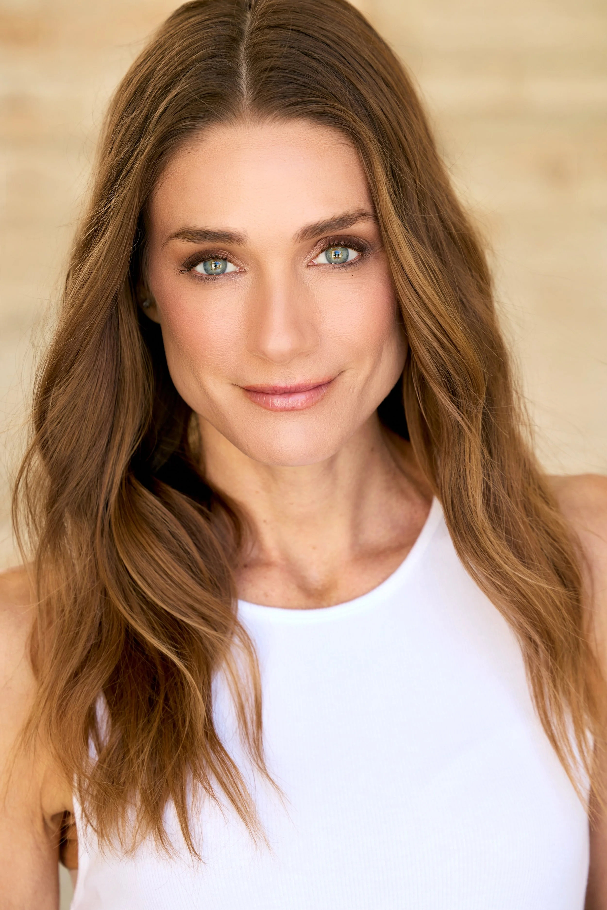 A woman with long, wavy brown hair and bright blue eyes smiling in a photo taken from her shoulders up, wearing a white top.