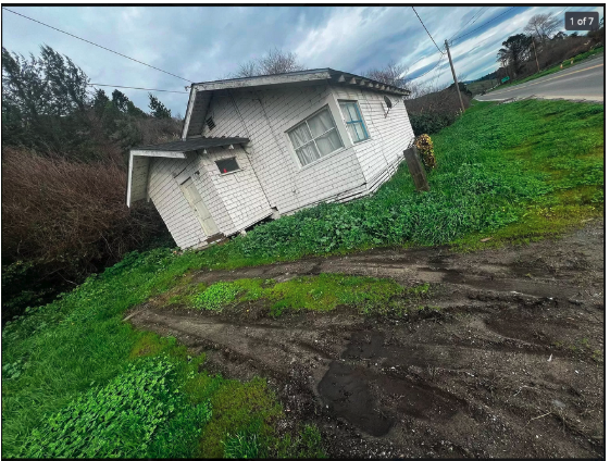 Tilted white house with windows, grass, dirt, and a road in the background under a cloudy sky.