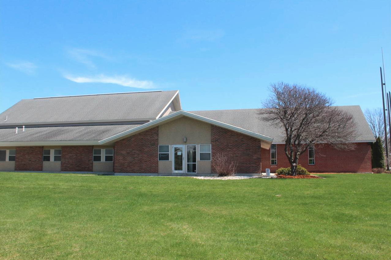 A single-story building with a combination of brick and beige exterior walls, set against a clear blue sky. It has a gabled roof, a glass front door, and several windows. There is a manicured lawn with a small tree and some shrubbery in front of the building.