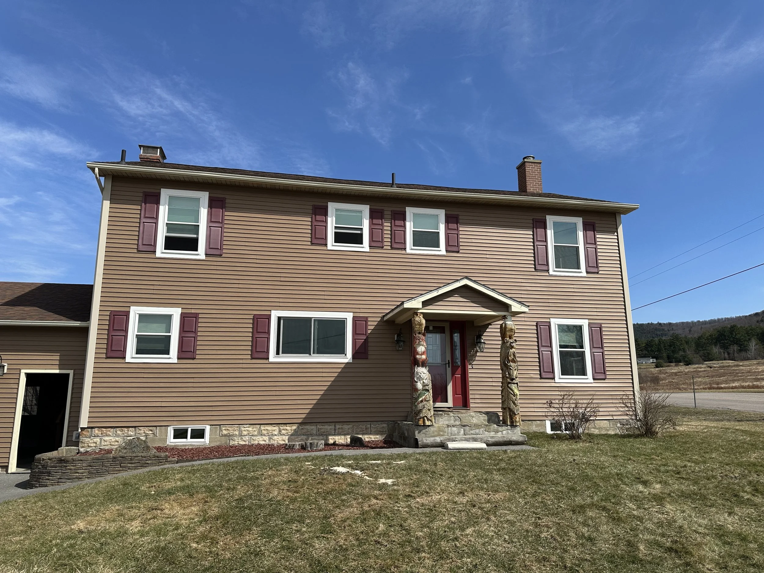 A three-story house with horizontal beige siding, purple shutters, a red front door, stone steps, stone foundation, and a small front yard, with a blue sky and some clouds in the background.