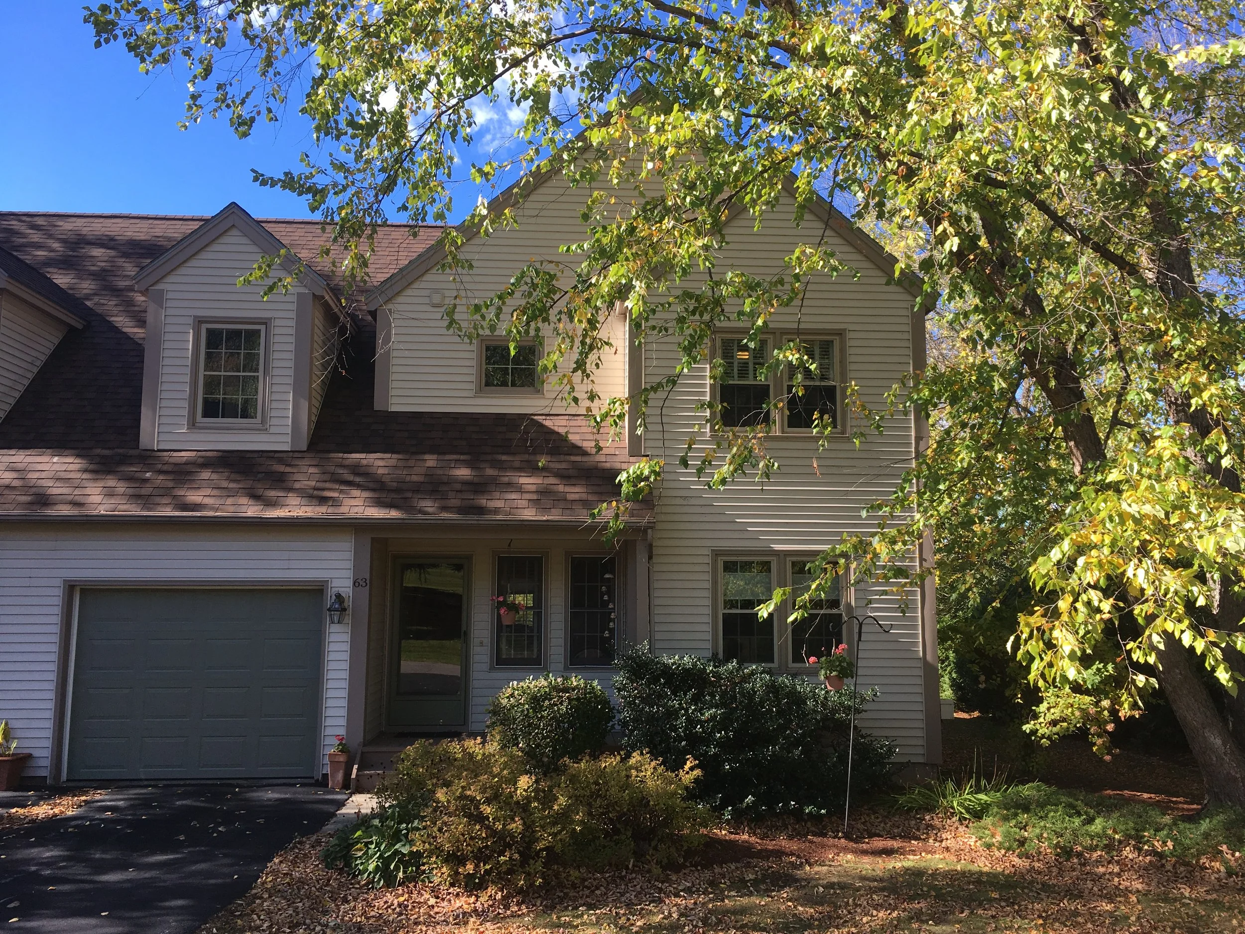 A two-story house with beige siding, brown shingle roof, and several windows, with front yard landscaping, trees, and a driveway.