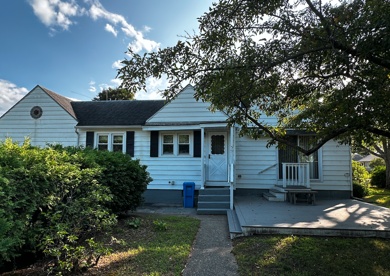 Front view of a light blue house with a small porch, steps, and a large tree shading the yard, under a partly cloudy sky.