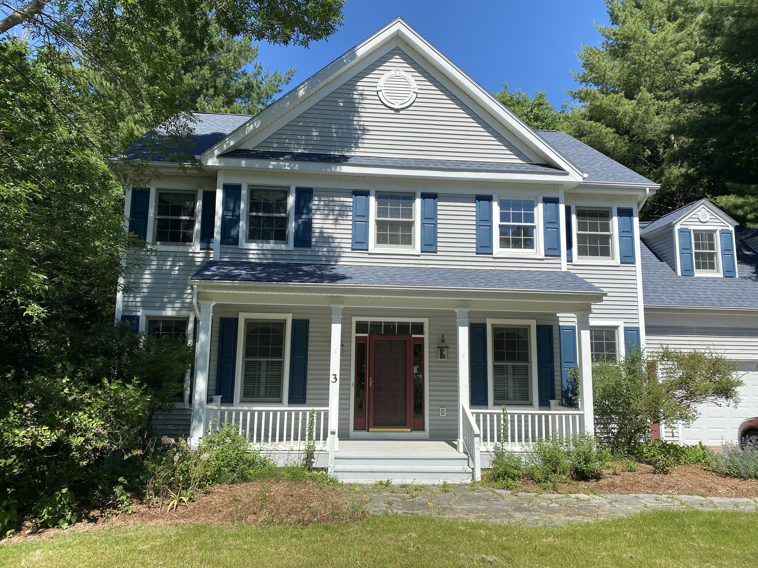 A two-story house with light gray siding and blue shutters, a front porch with white railing, a red front door, and surrounded by greenery and trees under a blue sky.