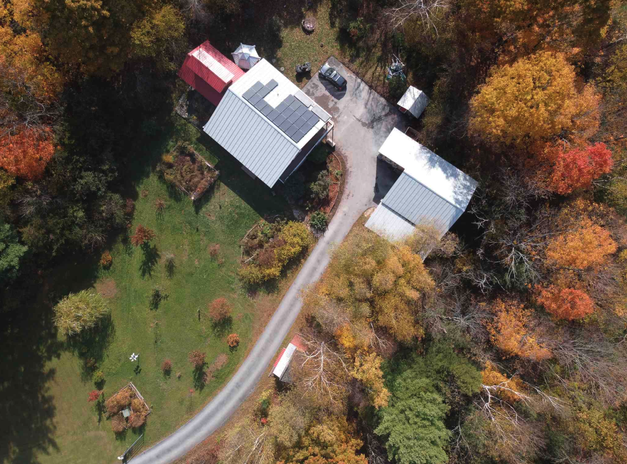 Aerial view of a property with two large metal-roof buildings, a driveway, a parked vehicle, trees with autumn leaves, and small garden areas.