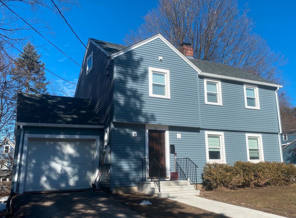 A two-story blue house with white trim, six windows, a front door, a one-car garage, and a rooftop chimney. There are leafless trees in the background and a sidewalk in front.
