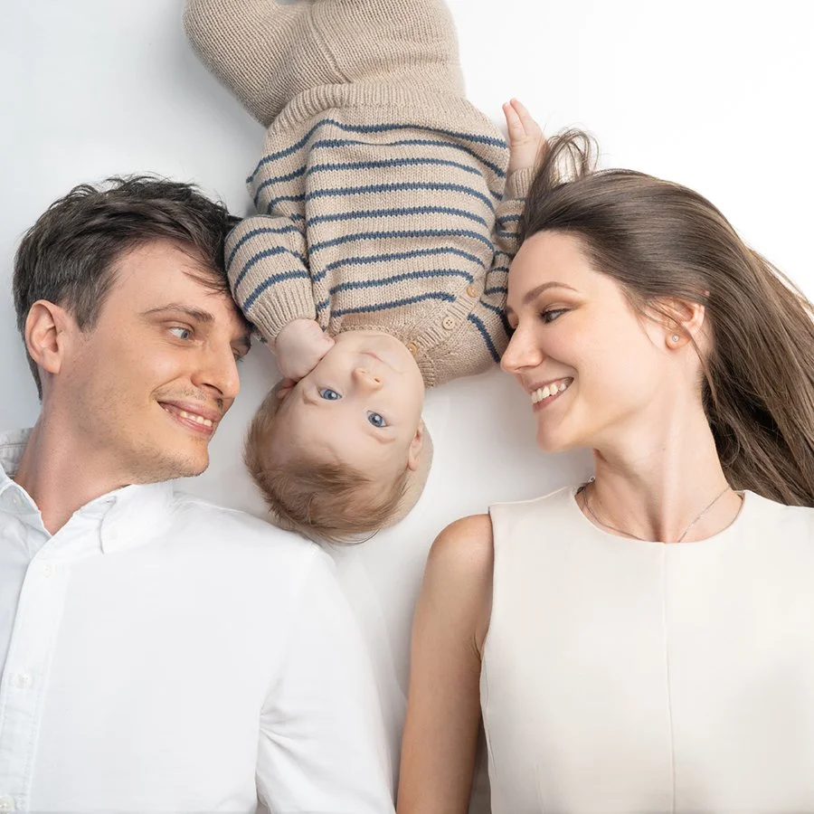 Familie mit Baby liegt auf weißem Hintergrund, blickt sich liebevoll an, Mutter und Vater lächeln.