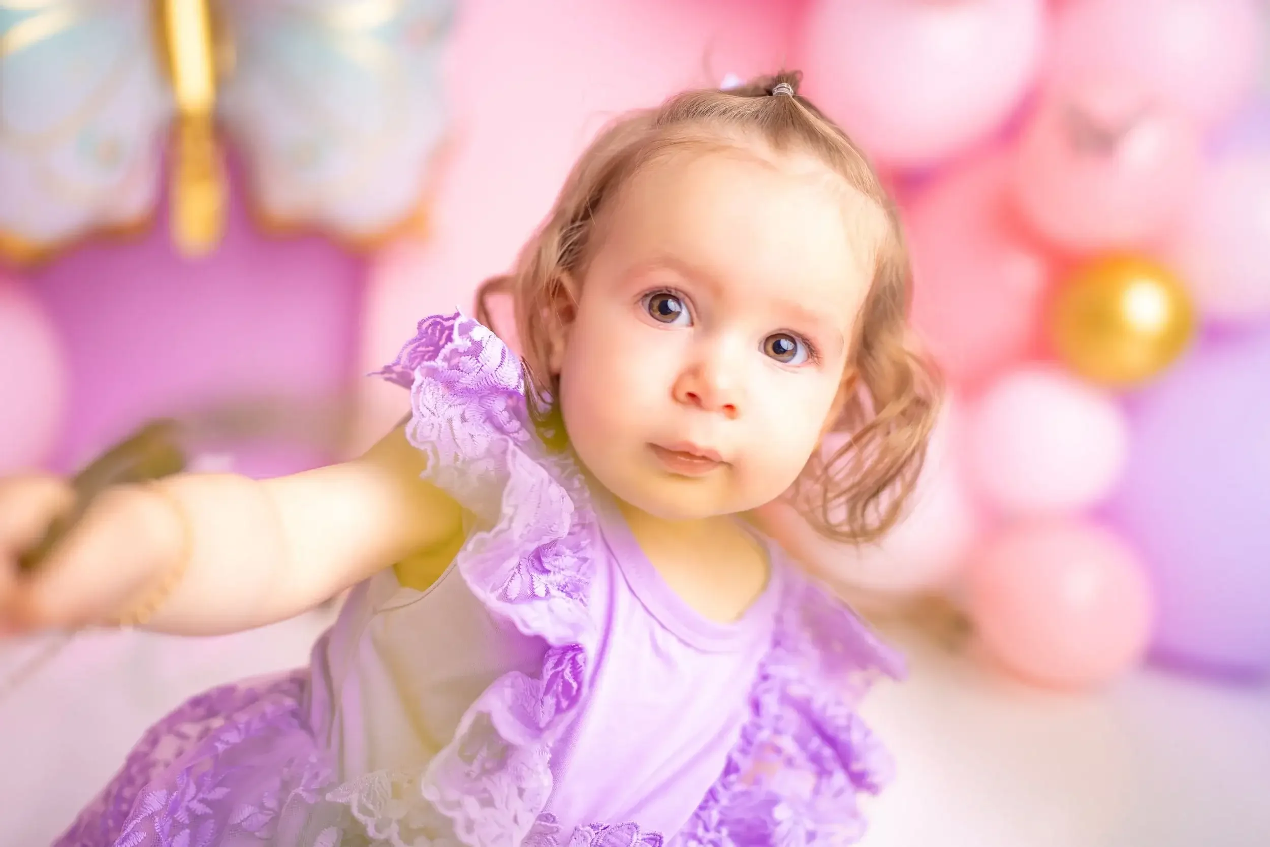 Mädchen im lila Kleid blickt vor einer rosa Ballonkulisse in die Kamera.