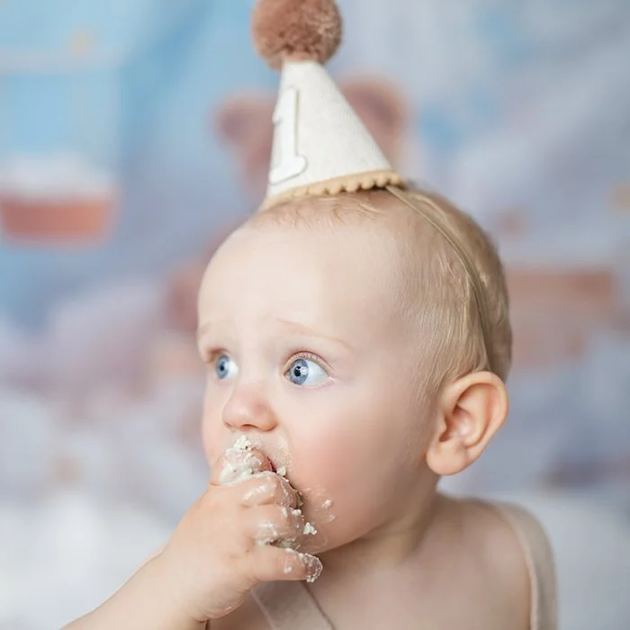 Ein Baby mit Partyhut isst Kuchen, Blick nach rechts, mit blauen Augen.