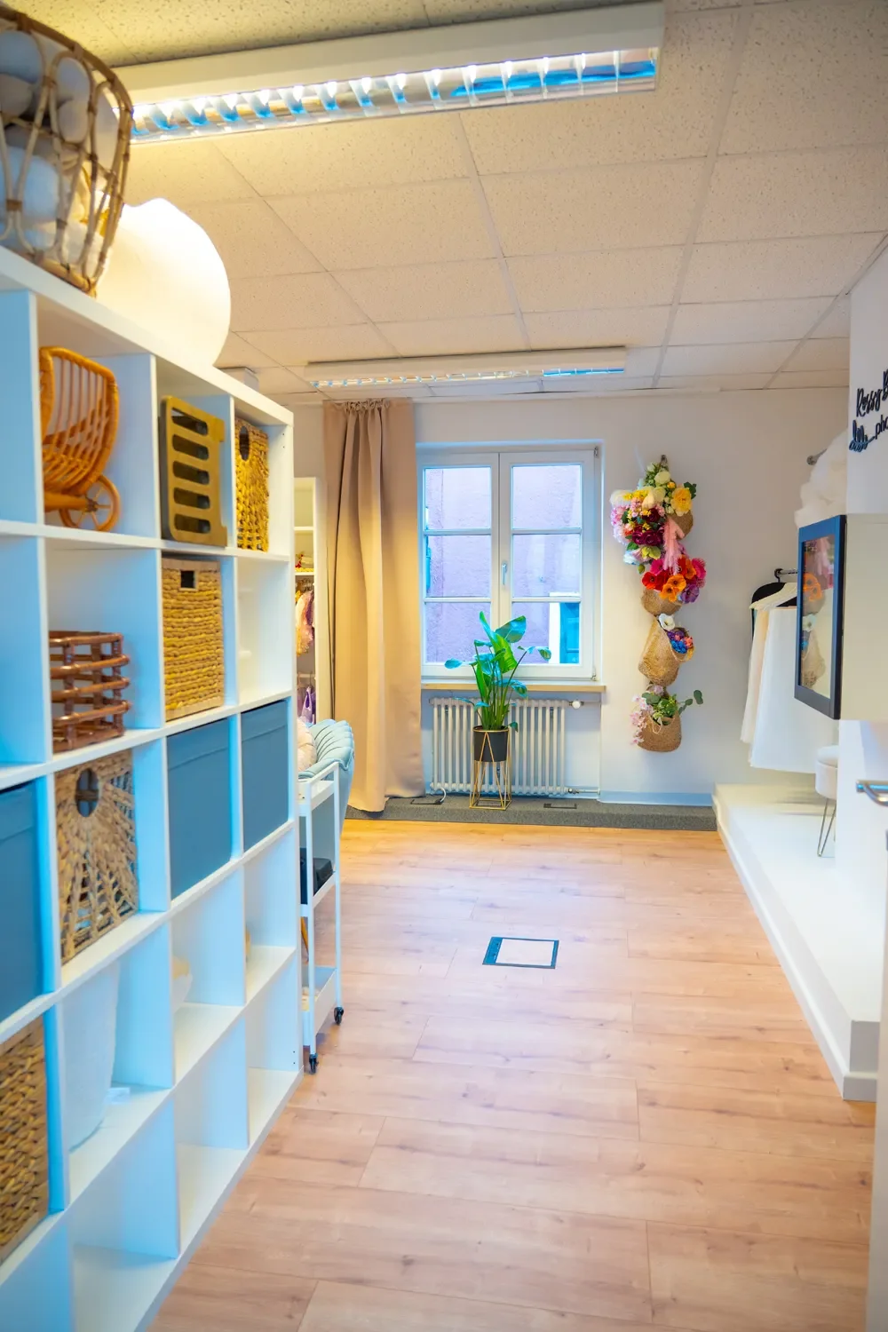 Ein gemütlich eingerichteter Raum mit einem Fenster, Pflanzen, gestrickten Wanddekorationen und Regalen mit Körben und Dekorationsgegenständen.
