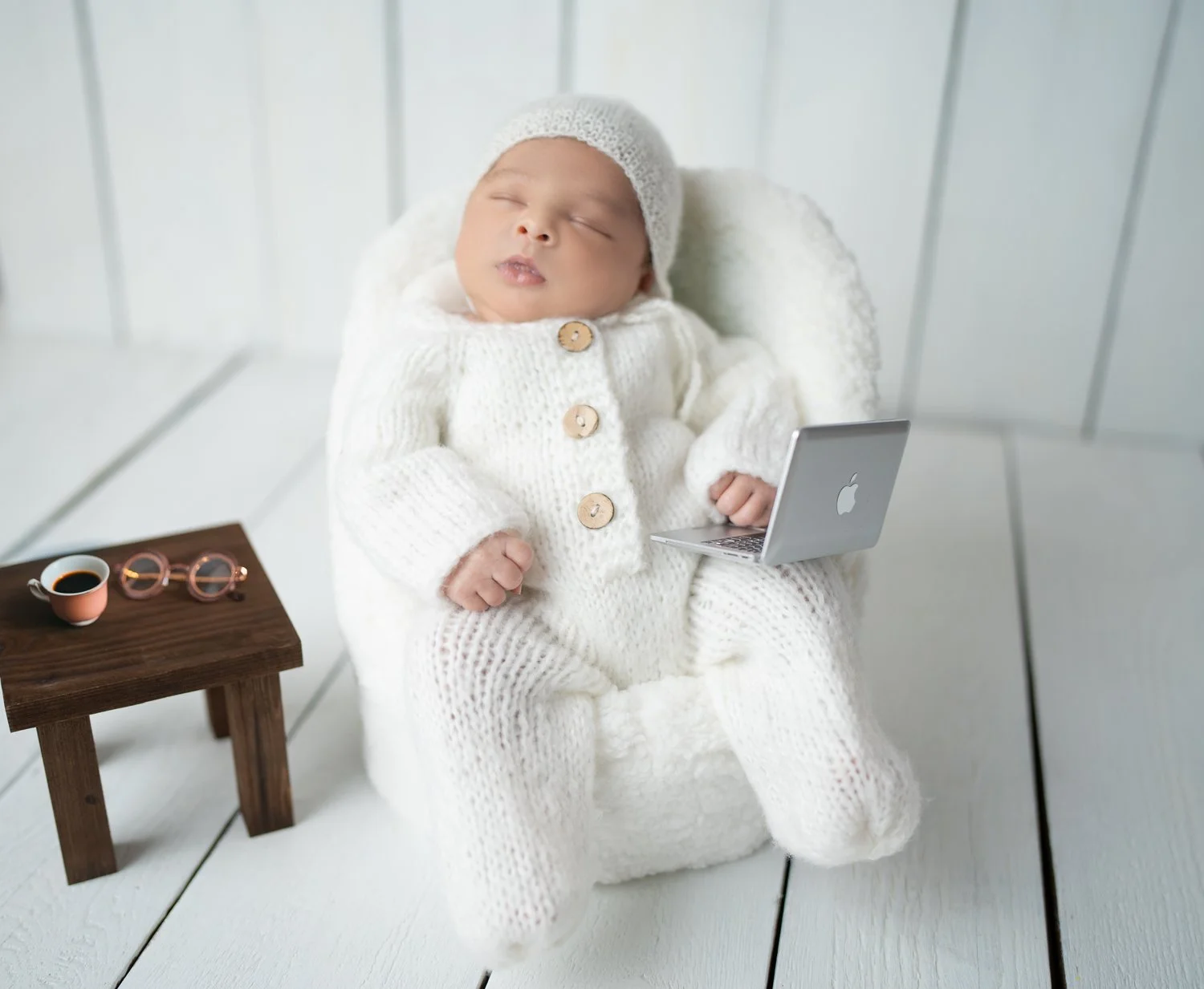 Ein schlafender Säugling in einem weißen Strickoutfit mit Mütze sitzt in einem weißen Sitz.