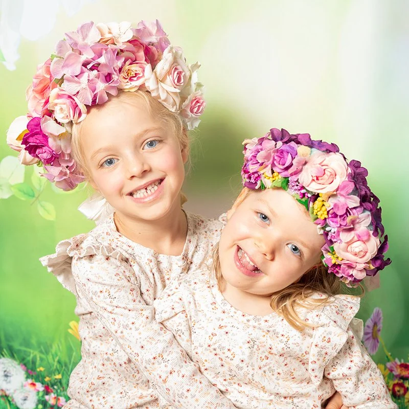 Zwei lachende Kinder, beide tragen Blumenkränze auf dem Kopf, vor einem grünen, blumigen Hintergrund.