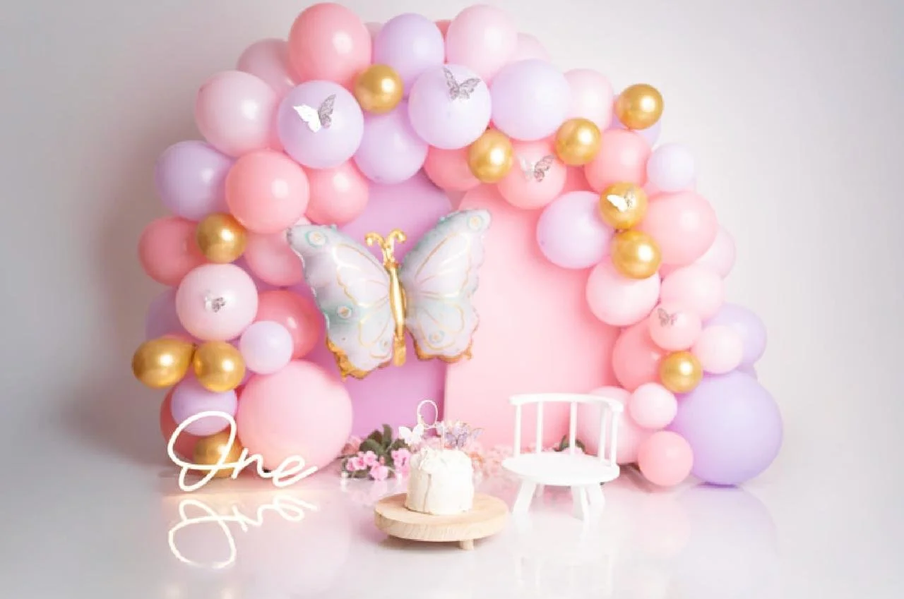 Cake Smash Dekoration mit vielen rosa, lila und goldenen Ballons, eine große Schmetterlingsfigur, ein weißer Gartenstuhl, eine kleine weiße Torte.