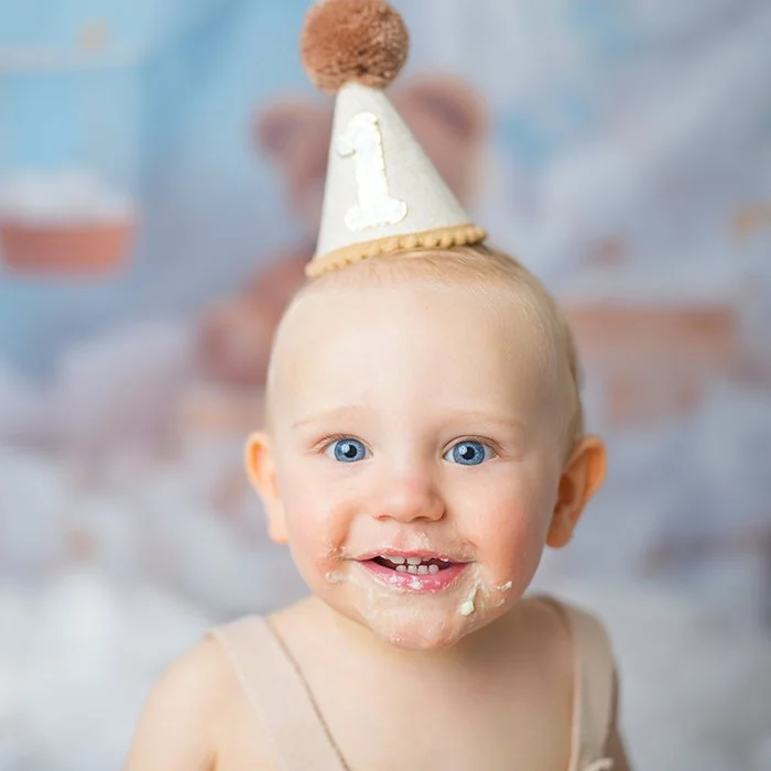 Ein Baby mit blauen Augen trägt eine Partyhaube mit der Zahl '1' und ist beim Essen, mit Kekskrümeln im Gesicht.