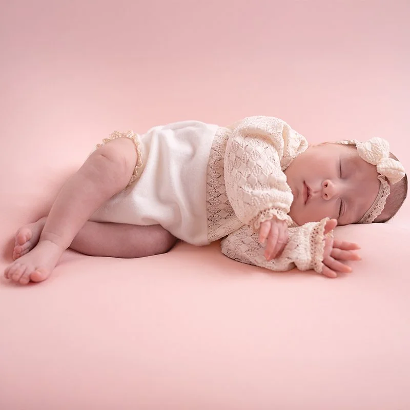 Ein schlafendes Baby liegt auf einem pinken Hintergrund, trägt ein weißes Kleid und eine gestrickte Mütze mit Schleife.