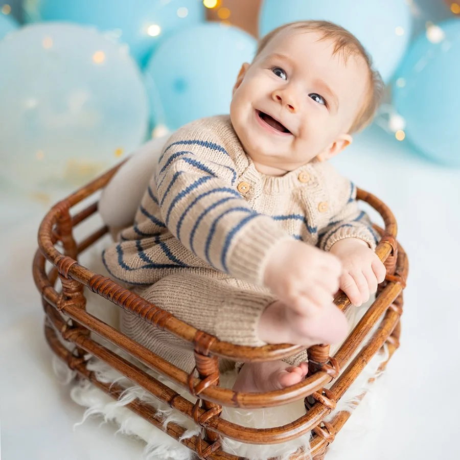 Lächelnder Säugling, der in einer kleinen Holzkorb sitzt, umgeben von blauen Ballons, bei einem Fotoshooting.