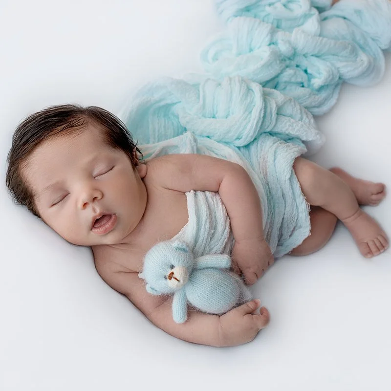 Neugeborenes Baby schläft, hält einen kleinen gestrickten Teddy, liegt auf weißen Bettunterlage, umhüllt von einer hellblauen Decke.