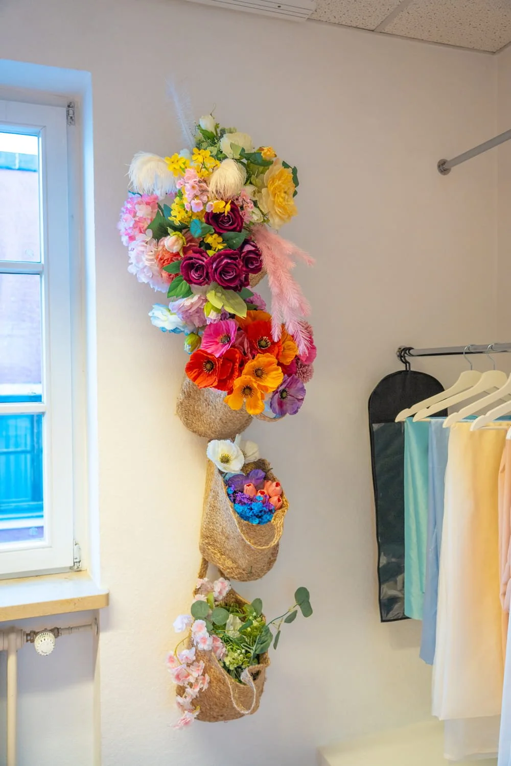 Ein vertikaler Wandkorb mit bunten künstlichen Blumen in einem Raum neben einem Fenster und Kleiderstangen.