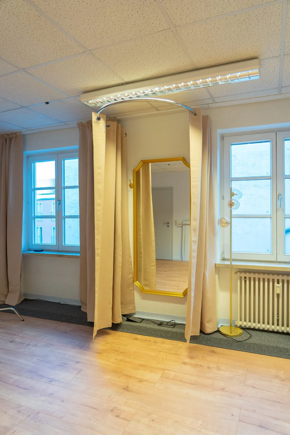 Ein Raum mit zwei Fenstern, einem Spiegel, einem Stehlampe mit mehreren Glühbirnen, Vorhängen und einem Holzfußboden.