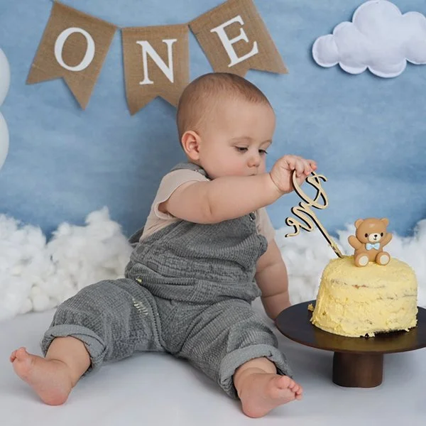 Ein Baby sitzt vor einer Torte mit einem Teddy auf einem Teller und hält eine Dekoration mit dem Wort 'One'.