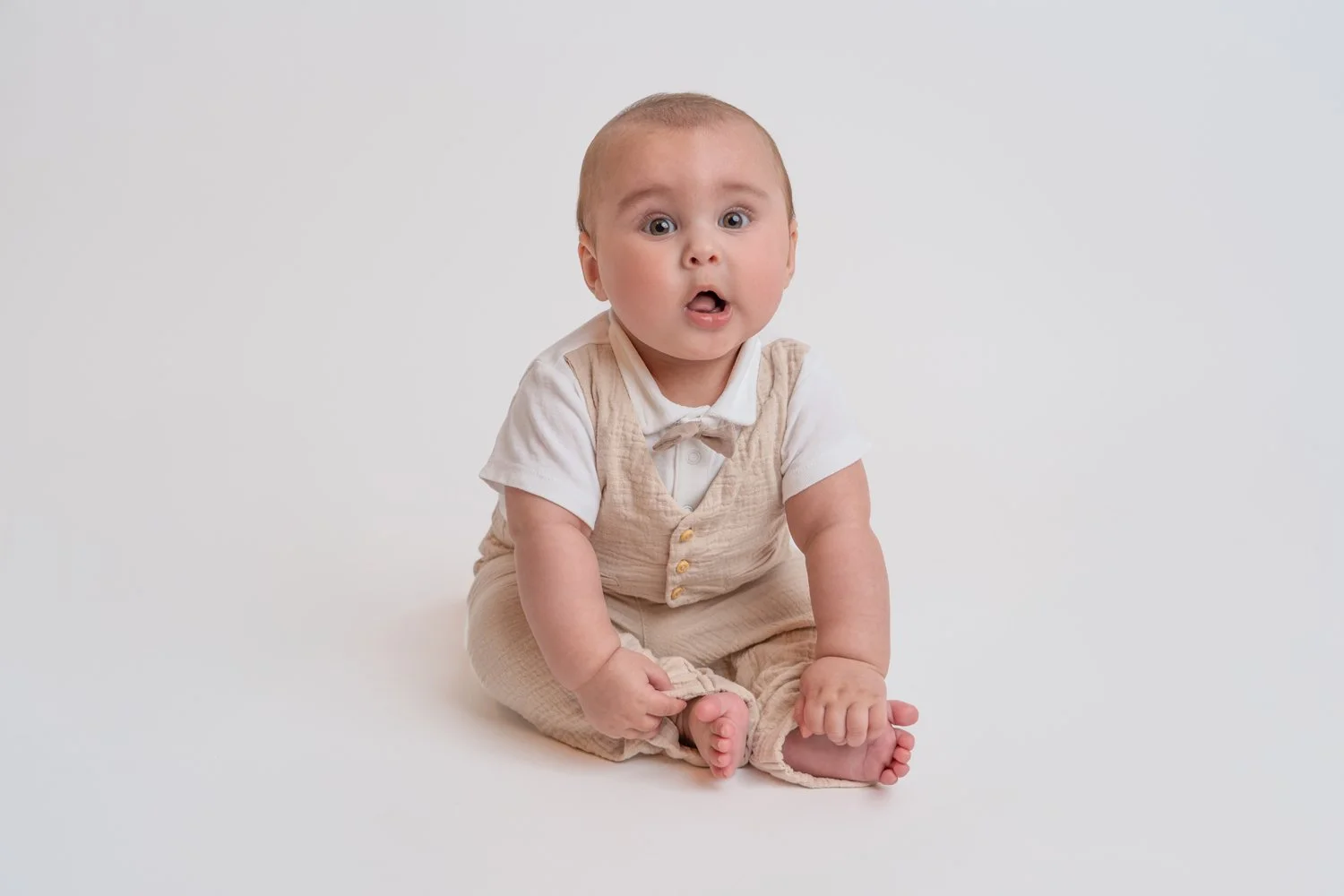 Ein Baby in beige Kleidung auf einem weißen Hintergrund, das überrascht schaut.
