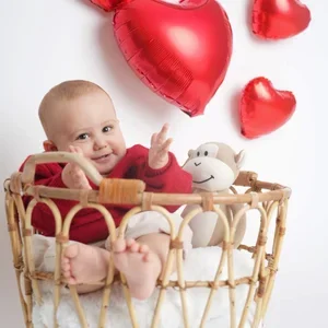Lächelndes Baby sitzt in einem Korb vor roten Herzballons.