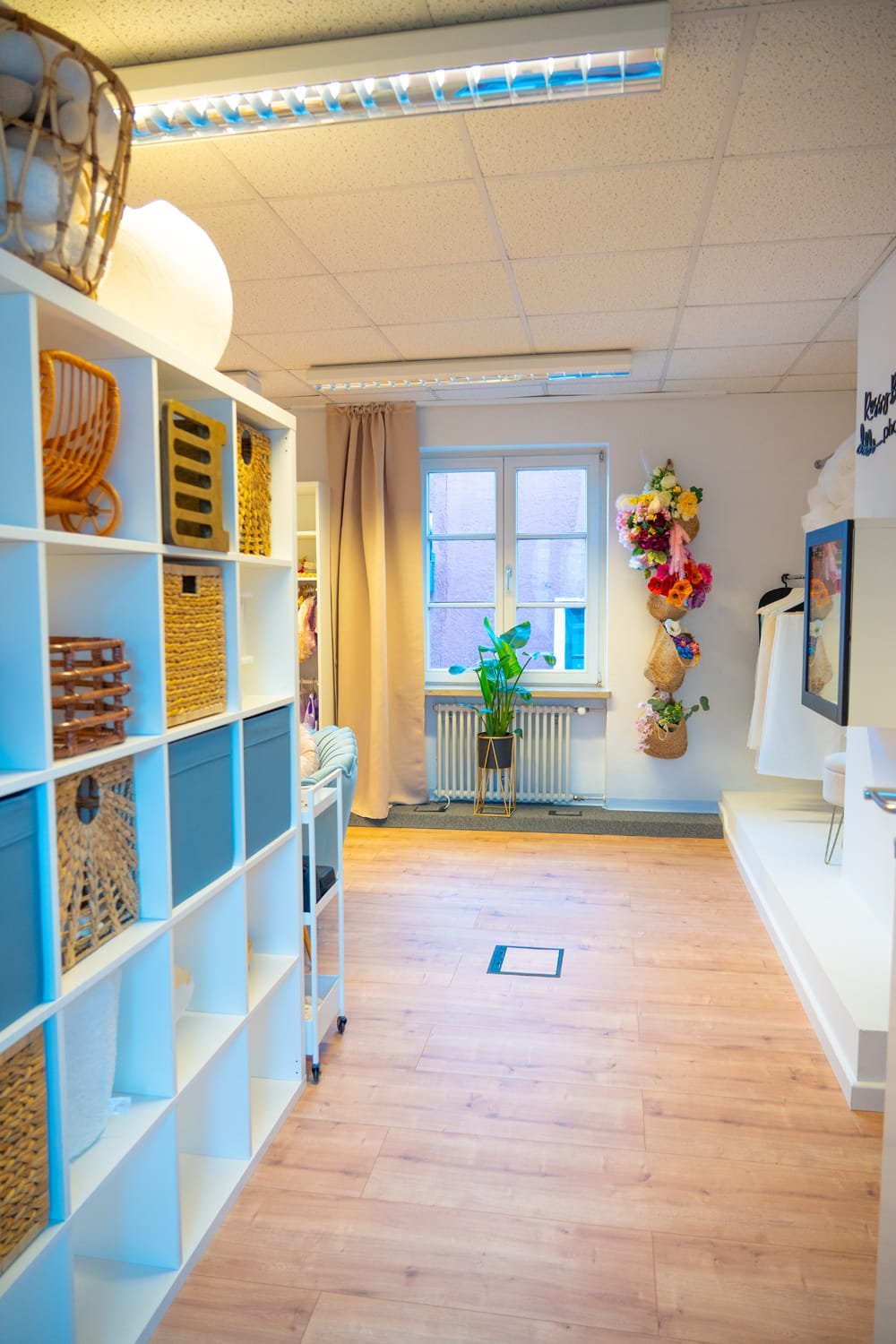 Ein hell erleuchteter Raum mit weißen Regalen, einem Fenster mit beige Vorhängen, einer Topfpflanze, einem Wanddekoration mit bunten Blumen und einem weißen Sitzbereich.