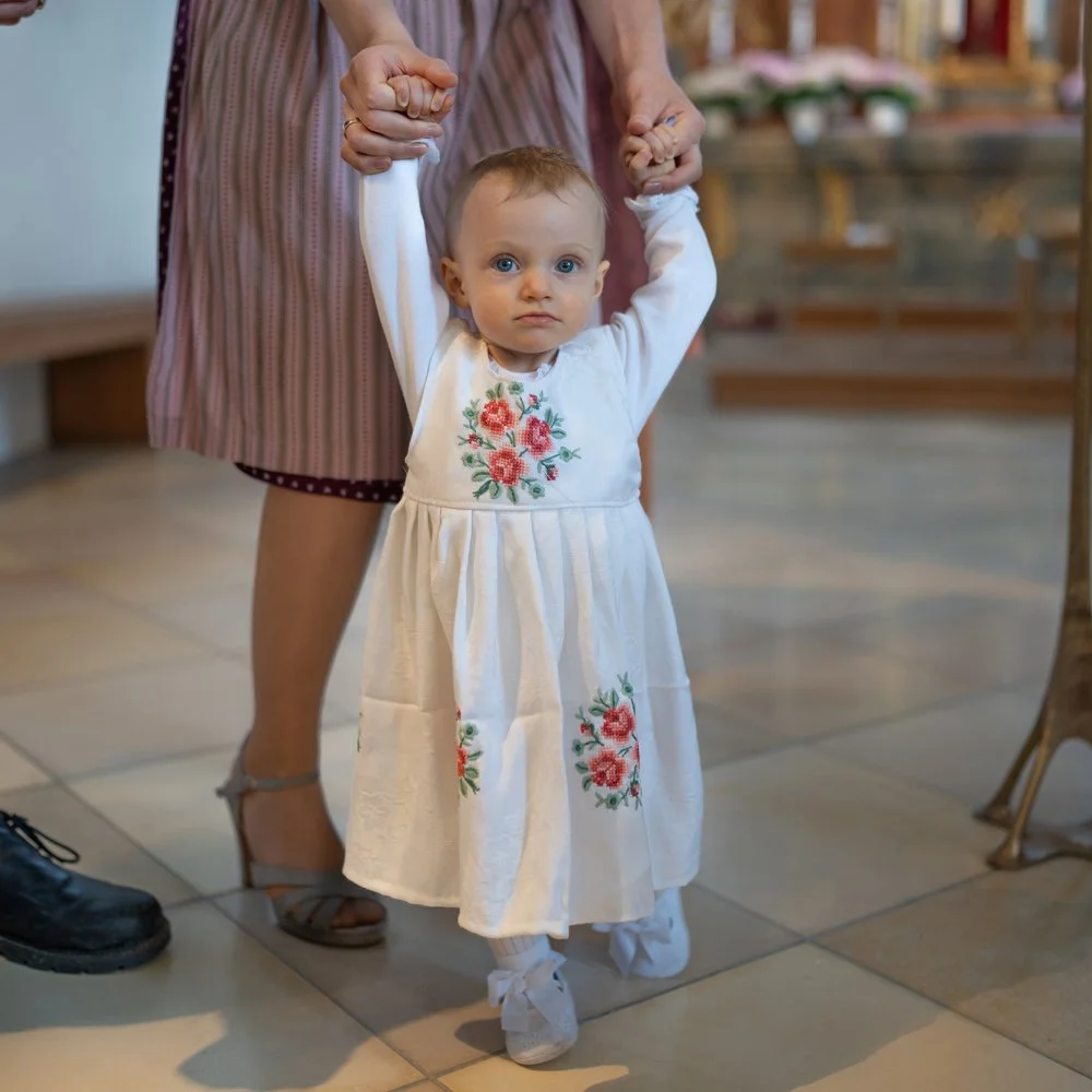 Ein kleines Mädchen mit blauen Augen trägt ein weißes Kleid mit floralem Muster und wird von einer erwachsenen Person an den Händen gehalten.