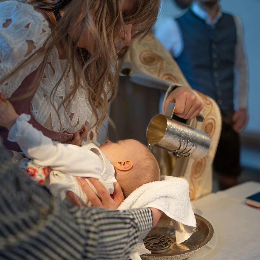 Ein Baby erhält eine Taufzeremonie, während eine Frau es hält und ein Priester Wasser auf den Kopf des Babys gießt.